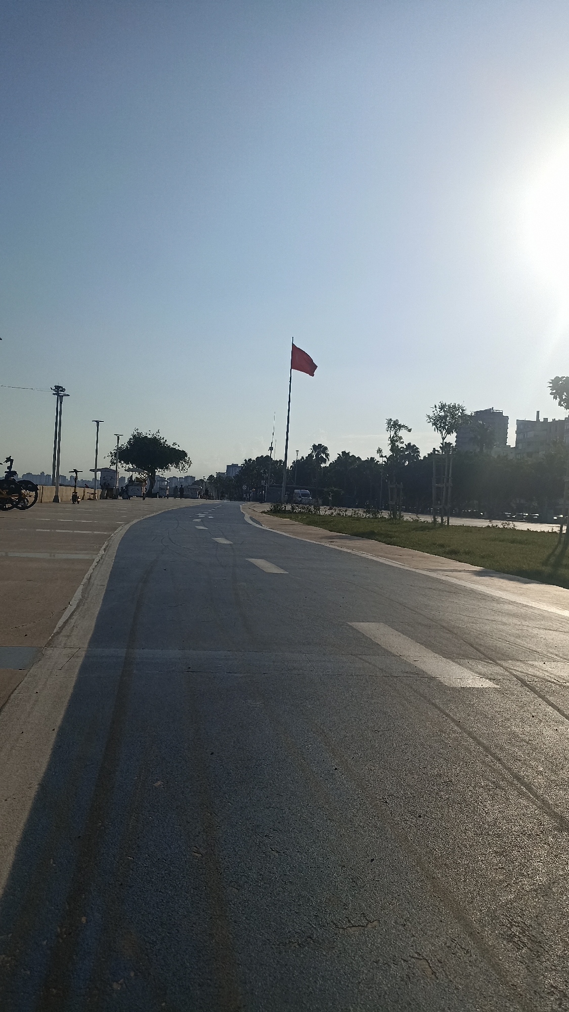 An exterior, eye-level, wide shot captures a paved road curving to the right under a clear, bright sky with the sun setting on the right. The road, marked with white dashed lines and arrow indicators, appears to be a pathway for vehicles or cyclists. To the left of the road is a concrete walkway with a shadow cast from an unseen object. A red flag flies from a tall flagpole in the middle distance. Beyond the walkway and road, a line of trees and buildings forms the urban landscape, with the city of Yenişehir, Turkey, serving as the backdrop. The scene is illuminated by the setting sun, casting long shadows and creating a warm, golden glow.