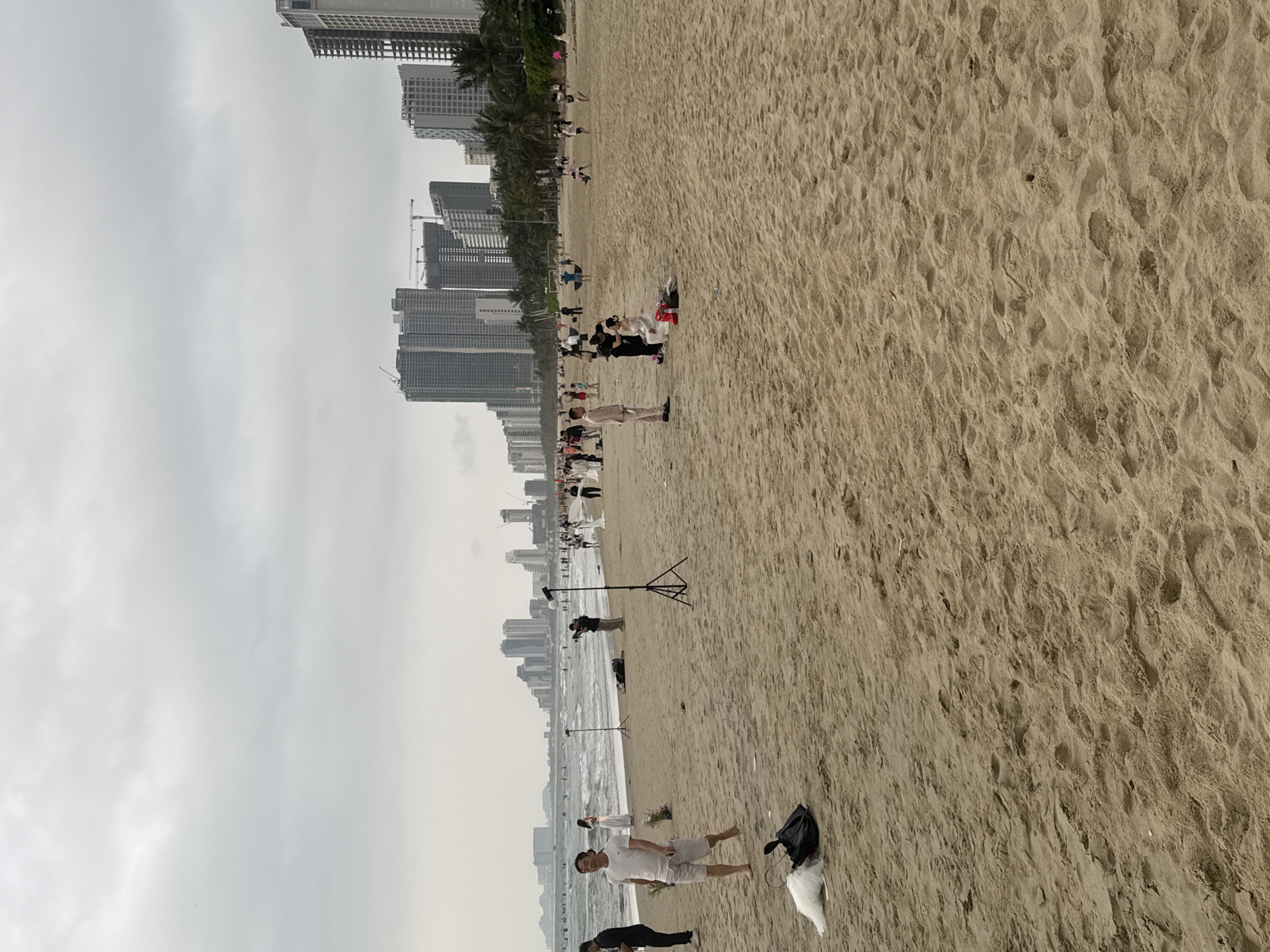 The image depicts a beach in Đà Nẵng, Vietnam. The beach is sandy with visible footprints. Numerous people are present, some walking along the shoreline, others gathered in groups. Several individuals appear to be involved in a photoshoot or filming session, with lighting equipment visible. In the background, a cityscape of tall buildings is prominent, defining the urban backdrop of the beach. The sky is overcast, suggesting a cloudy day.