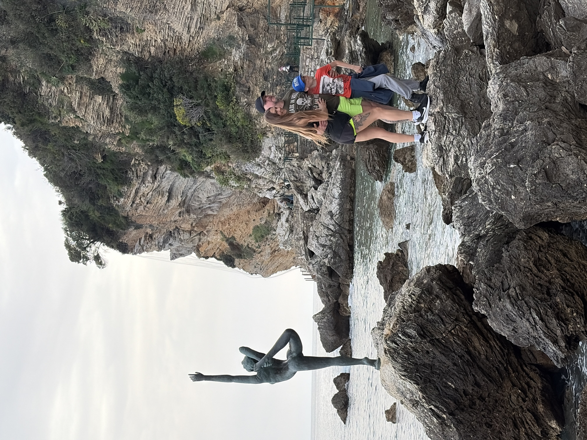 The image shows a group of three people and a statue in a rocky coastal environment. The statue, which appears to be bronze, depicts a woman in a yoga pose, extending one arm toward the sky. The statue is located in the water on some rocks. The people consist of a man wearing a hat, t-shirt, and pants; a woman wearing shorts and a t-shirt, hugging the man; and a young boy in a red t-shirt. All three are standing on rocks near the water. The environment includes a rocky shoreline and cliffs covered with vegetation. The sky is overcast. The location is Budva, Montenegro.