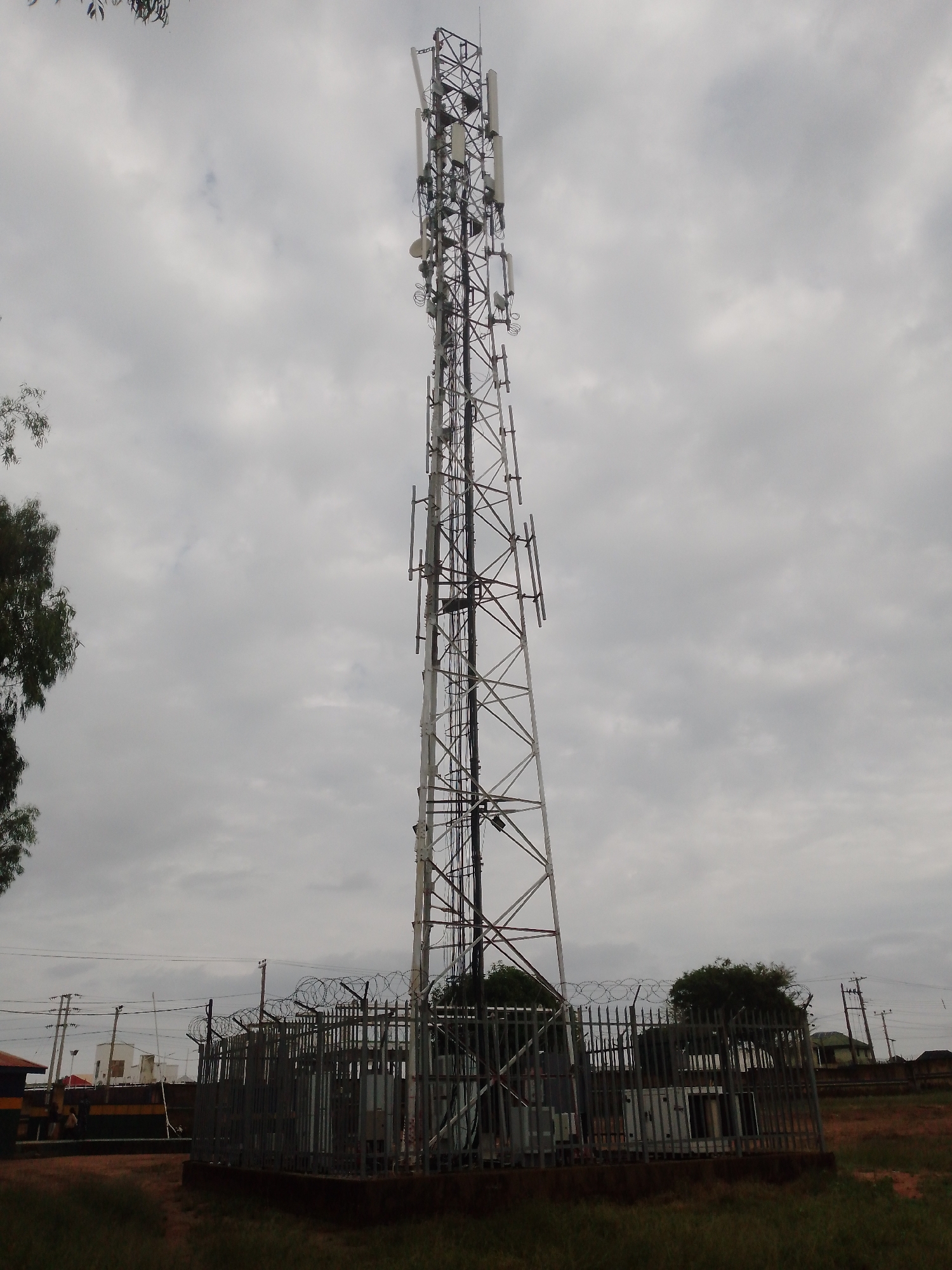 The image shows a tall telecommunications tower in Unguwar Saya, Nigeria. The tower is made of metal lattice and is equipped with several antennas. It is surrounded by a fence with barbed wire. In the background, there are power lines and buildings, including houses and what appears to be a small commercial structure. Trees are also visible in the scene. The sky is overcast, with grey clouds. No people are clearly visible in the image. The ground is covered with grass and some dry earth. The scene depicts a typical telecommunications infrastructure setup in a semi-urban or rural area of Nigeria.