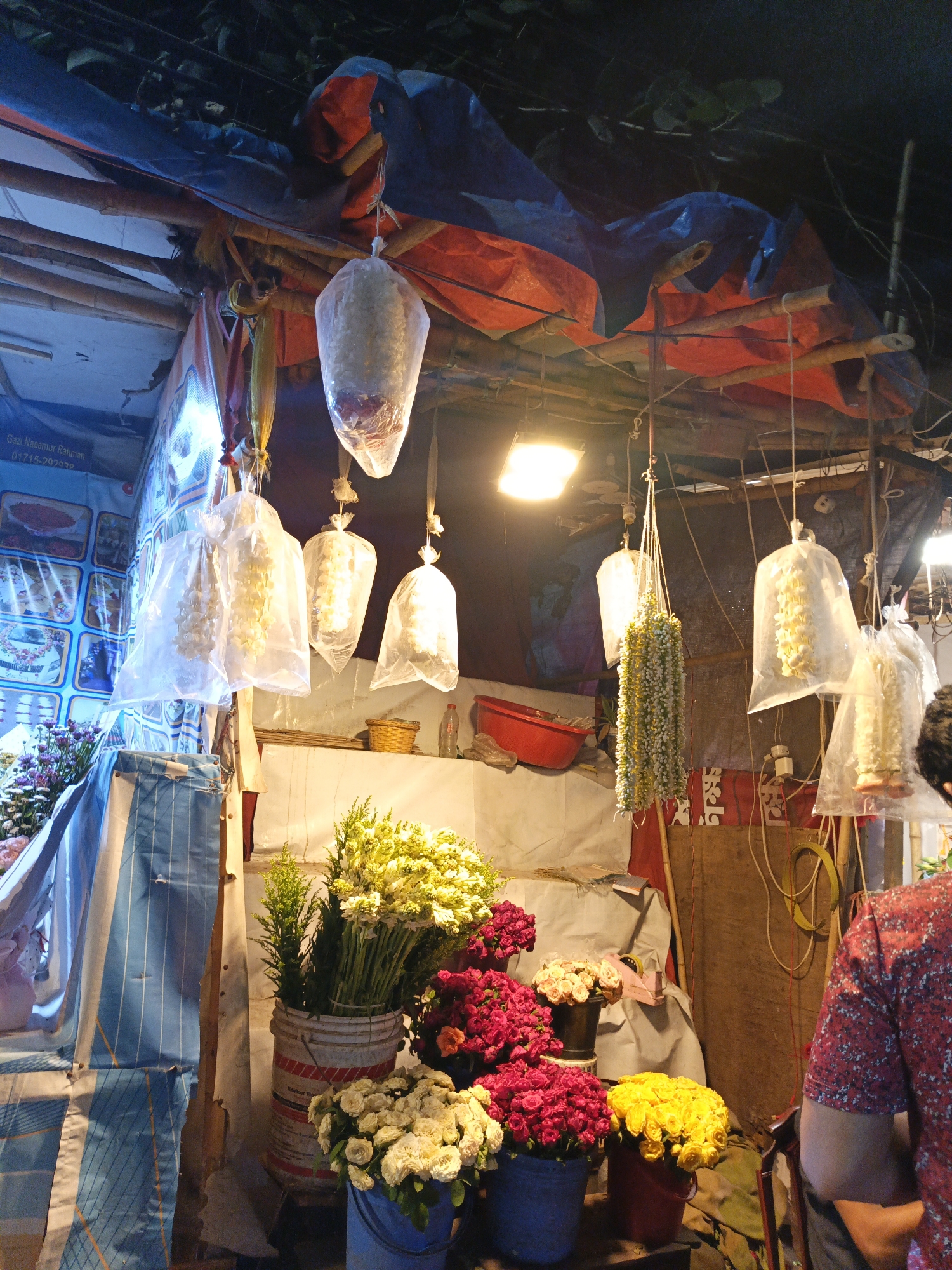 This is a nighttime scene depicting a flower shop in Dhaka, Bangladesh. The shop is under a makeshift shelter constructed of wood and draped with blue and orange tarps. Hanging from the structure are multiple clear plastic bags, each containing flower garlands, suggesting these are for sale. Buckets filled with various flowers including yellow, red, and white roses, as well as yellow spray flowers are arranged at the front of the shop. A bucket with green foliage is also seen. A man in a red floral patterned shirt is partially visible on the right side of the image, presumably a customer. In the background, behind the flowers, there is a white wall with some decorations and objects. One can also see the corner of a menu with images of food, adding to the atmosphere of a busy street. Artificial lighting illuminates the scene.