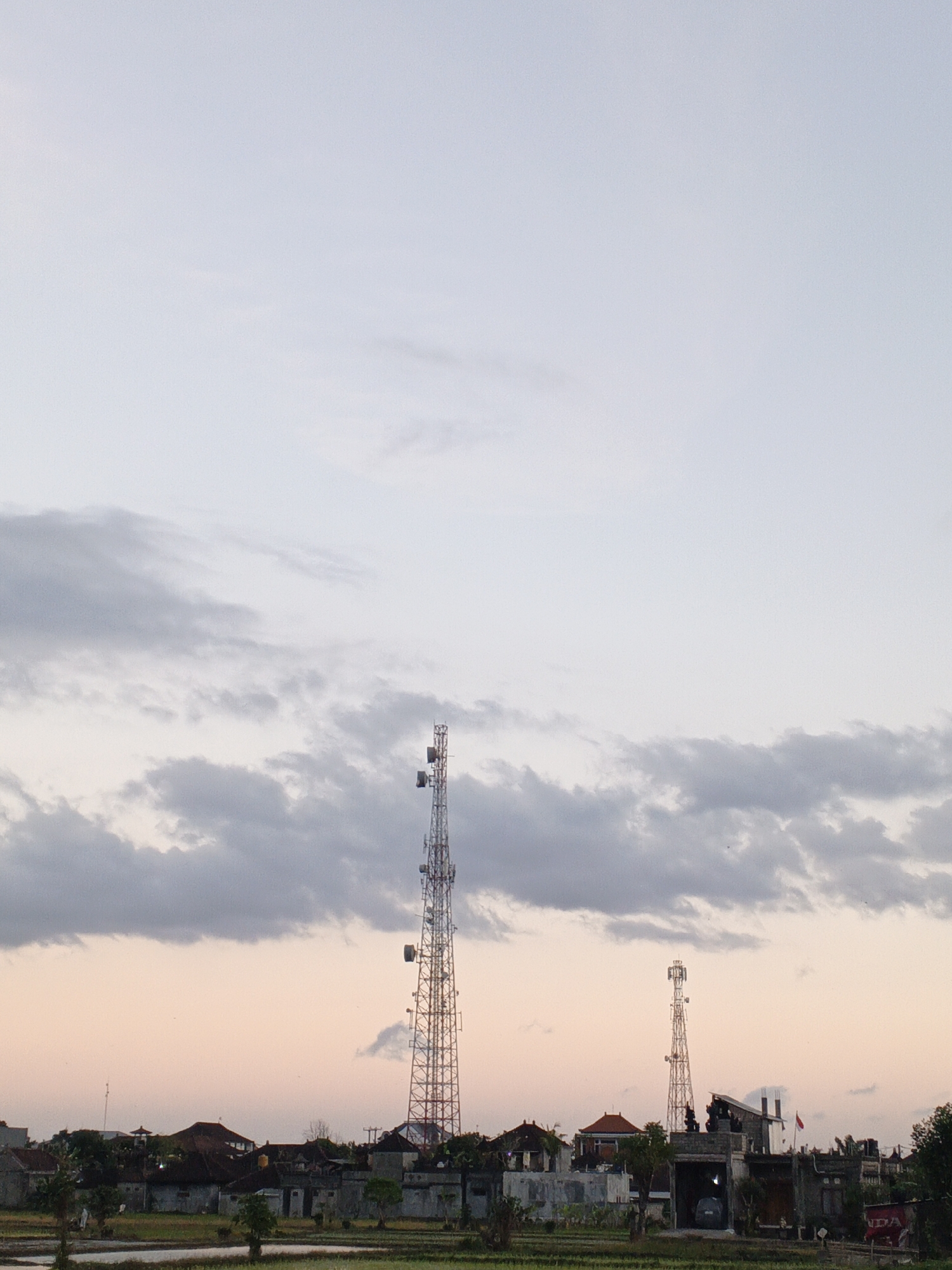 The image depicts a landscape view in Sukawati, Indonesia, at what appears to be dusk. Two tall telecommunication towers dominate the mid-ground, with the larger one on the left and a slightly shorter one to its right. Below the towers, a cluster of buildings with traditional Balinese roofs are visible, interspersed with trees and vegetation. In the foreground, what seems to be a rice paddy or agricultural field with water can be seen. The sky above is a gradient of pale blue and soft pink, with scattered, wispy grey clouds. The overall atmosphere is tranquil and serene, capturing the transition from day to night in a rural Indonesian setting.