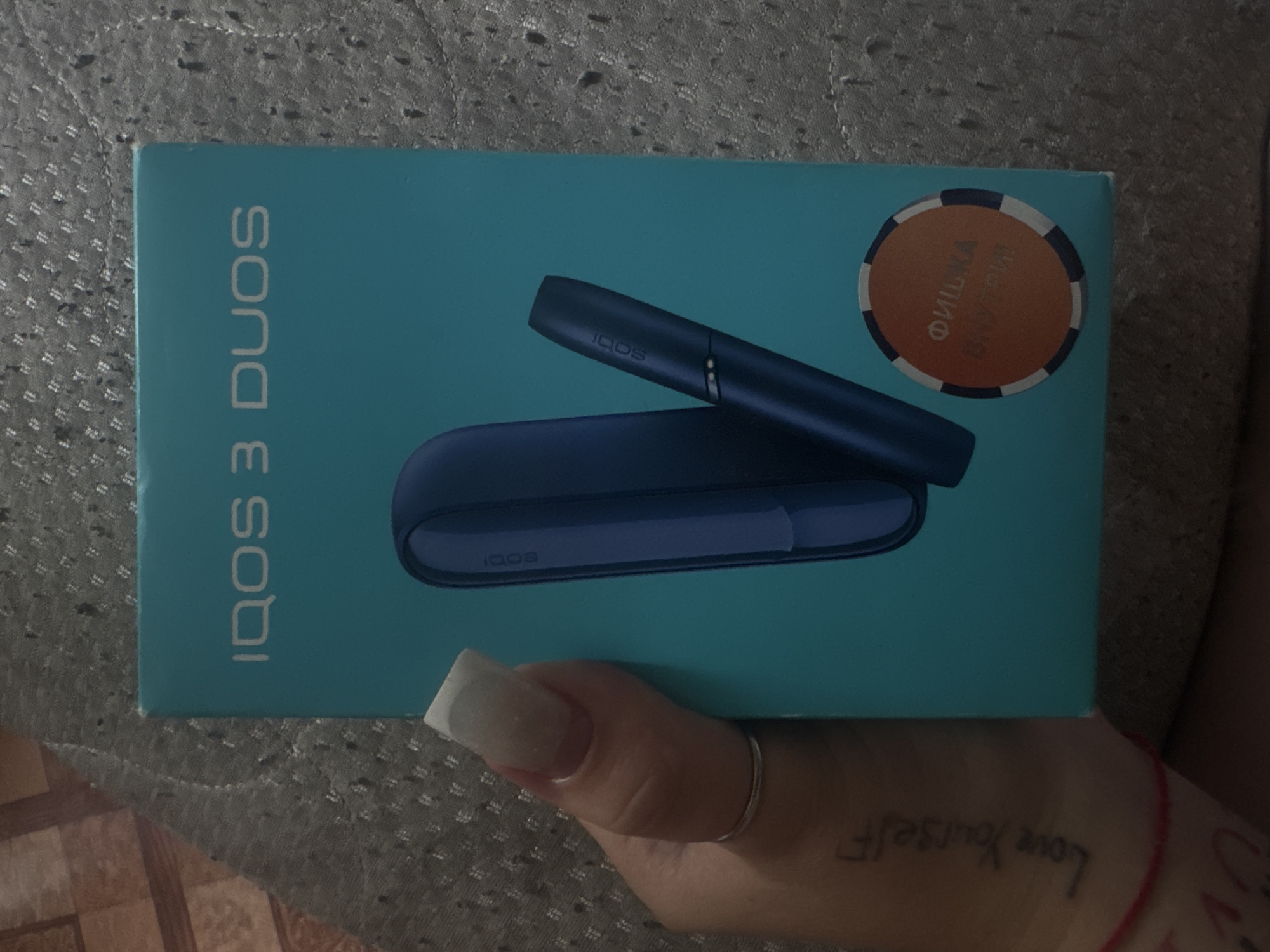 The image shows a hand holding an IQOS 3 DUOS product box. The box is blue and features the IQOS logo and "3 DUOS" text. Inside the box are two blue IQOS devices, a holder and a pocket charger. The hand holding the box has long, white-tipped fingernails and a silver ring on one finger. A tattoo reading "Love Yourself" is visible on the hand. The background is a textured gray surface, possibly a blanket or carpet. A circular orange sticker with Cyrillic text is on the lower right of the box. The scene is likely indoors, and the lighting suggests it could be daytime. There is no information in the image that places it specifically in the city of Razvedchik, Russia.