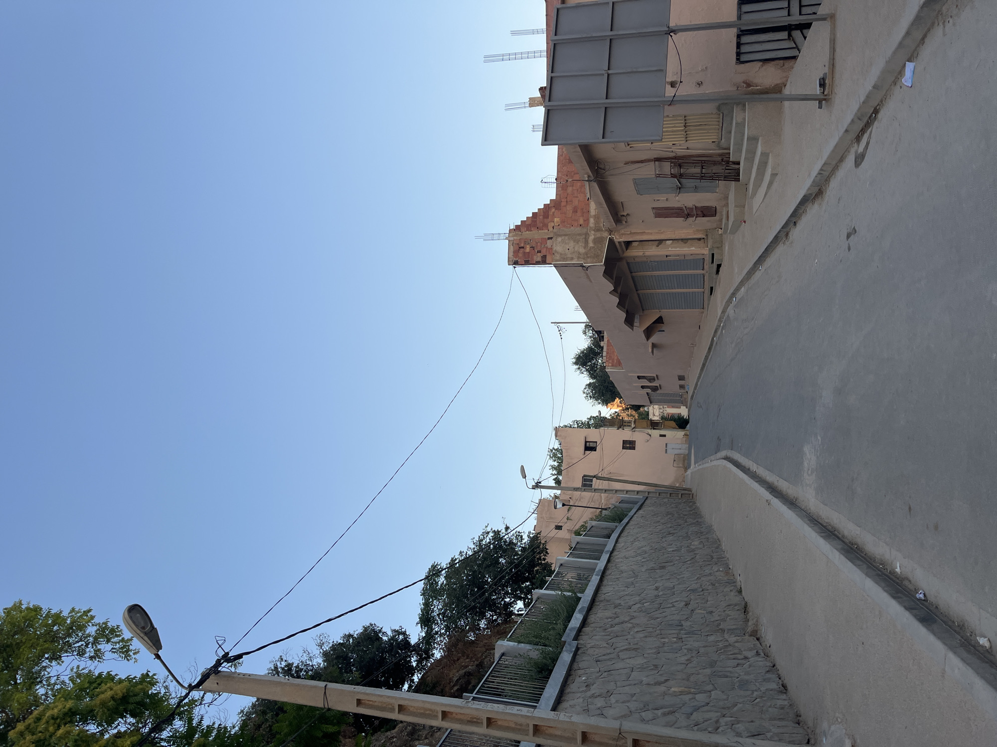 This is a photograph of a street in El Hajeb, Morocco, taken during the day. The street is paved with asphalt and slopes upwards into the distance. On the left side of the street, there is a tall, curved stone wall with a metal railing on top, behind which are trees and foliage. A concrete lamppost with a street light is visible near the top of the wall, connected by wires to other structures.
On the right side of the street, there are several buildings, mostly with pale pink facades. Some of these buildings appear to be shops with closed shutters. One building is under construction, with exposed brickwork and rebar sticking out from the top. There are also several steps leading up to the entrances of some buildings. A large, gray metal advertising board is mounted on a pole in front of one of the buildings. The sky is clear and blue. There are no people visible in the image. The scene depicts a quiet, residential or commercial street with a mix of older buildings and some under development.
