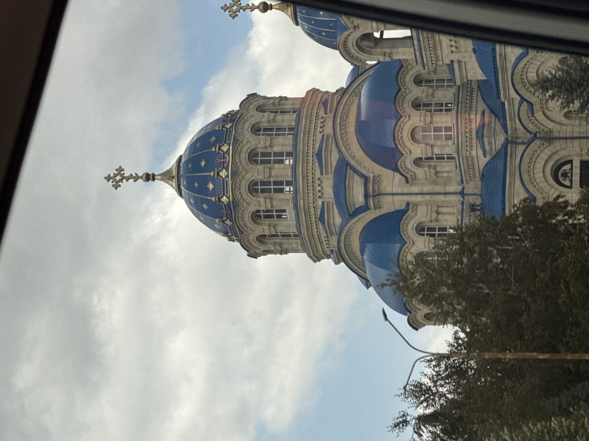 An exterior, eye-level shot captures a large, light-colored stone church with multiple blue domes topped with gold crosses. The dome structure is ornate with many arched windows. In the foreground, the top of a dark car windshield is visible, framing the scene. To the left, a tall, slender lamppost stands against a backdrop of dark green trees and a cloudy sky. The image appears to have been taken from inside a moving vehicle, suggested by the framing of the car and the slight motion blur.
