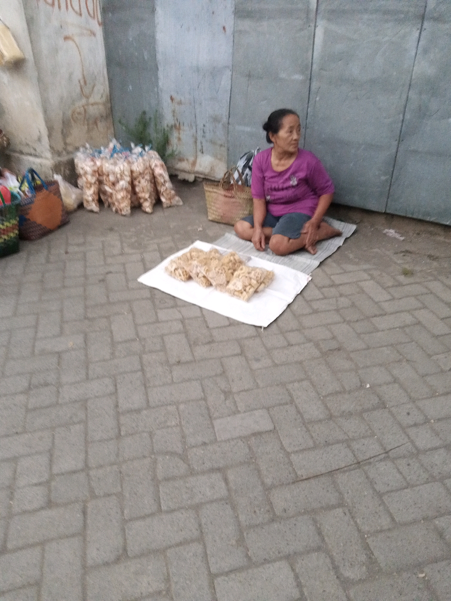 An outdoor, medium shot captures an elderly East Asian woman sitting cross-legged on a mat on a paved pathway in Sono Ageng, Indonesia. She is wearing a purple t-shirt and dark shorts, with her dark hair pulled back in a bun. In front of her, on a white sheet, are several clear plastic bags filled with what appear to be snacks. To her left, there are more bags of snacks, some woven baskets, and other shopping bags. The background features a weathered, gray wall or structure. The pathway is paved with rectangular grey bricks.