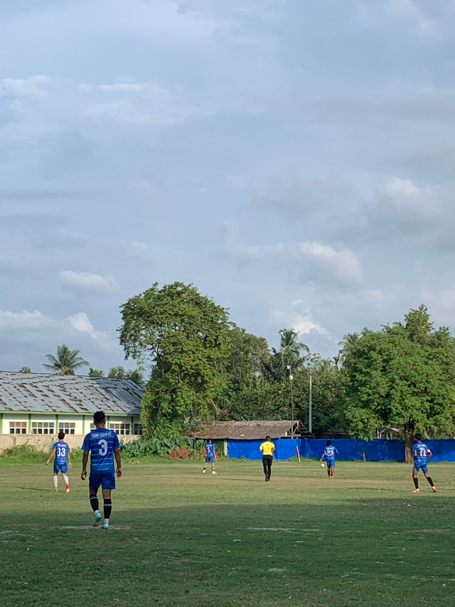 The image depicts an outdoor soccer game taking place on a grassy field in Keudelapang, Indonesia. The sky is partly cloudy. On the left side of the frame, a building with a corrugated metal roof and white walls with windows is visible. Several young males in blue soccer uniforms are scattered across the field. One player in the foreground, wearing number 3, is facing away from the camera and appears to be walking. Another player to his left, wearing number 13, is also in a blue uniform. Further in the background, there are other players in blue uniforms and one individual in a yellow shirt and black pants, potentially a referee or coach. Lush green trees and foliage surround the playing area, and a blue tarp or barrier is visible in the background. The overall scene suggests a casual soccer match or practice session.