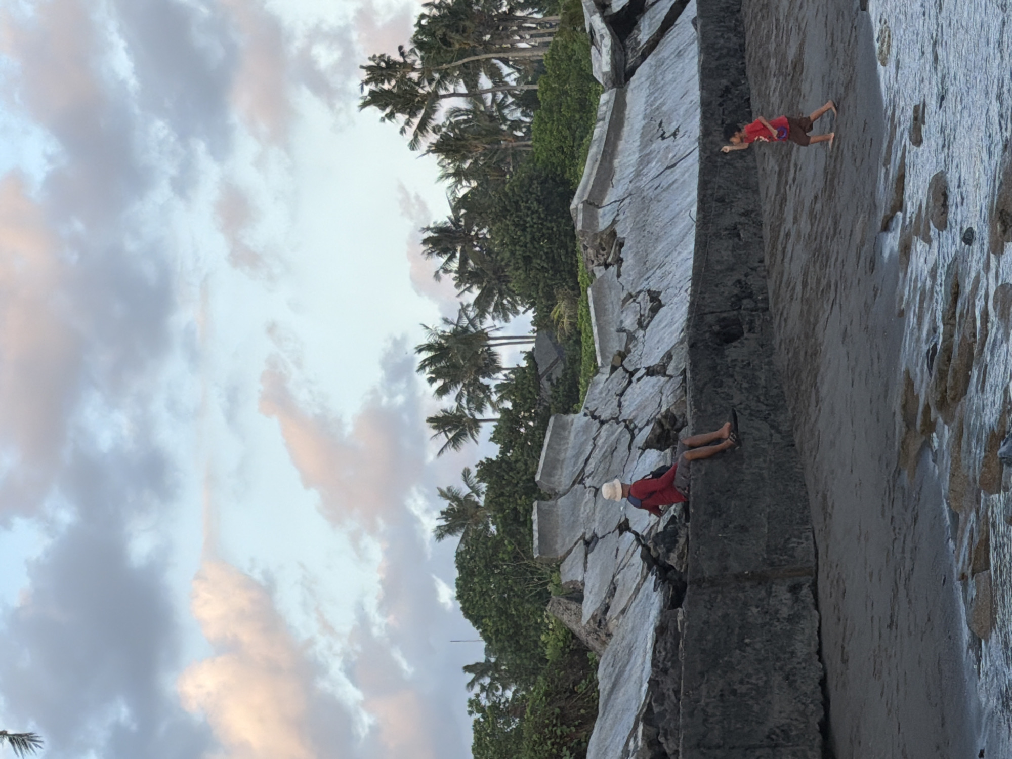 This image depicts a coastal scene in Mengwi, Indonesia, at what appears to be sunset or a cloudy day. In the foreground, a person wearing a red shirt and shorts sits on a concrete wall, facing away from the camera, with a straw hat on their head. To the right, a young child in a red shirt and brown shorts is running on the sandy beach, appearing to fly a kite, although the kite itself is not clearly visible. The background features a lush green landscape with numerous palm trees and dense foliage. A broken, concrete seawall is prominent in the midground, showing signs of significant damage and erosion. The sky is filled with soft, diffused clouds, tinged with hints of pink and orange, suggesting twilight. The ocean laps gently at the rocky shoreline in the lower portion of the frame.