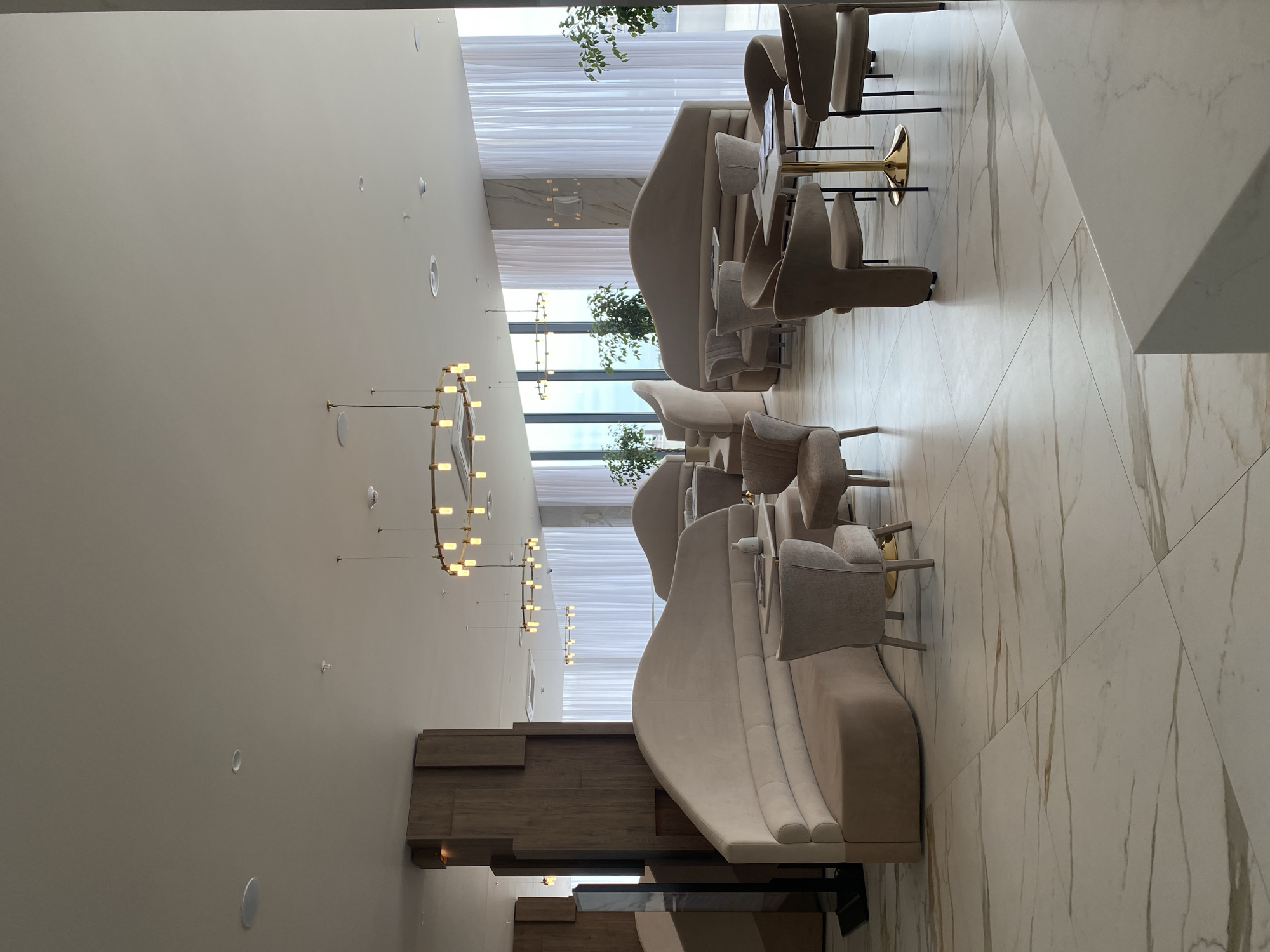 This is an interior shot of a lobby, likely in a hotel, possibly in Sochi, Russia, though no location indicators are visible in the image. The space is designed with a modern aesthetic, featuring a palette of neutral tones and clean lines.
The room is furnished with plush, curved couches in a light beige color. Matching armchairs are arranged around the couches. There are also small, gold-colored tables interspersed between the seating arrangements.
Lighting is provided by a series of circular chandeliers with exposed bulbs. The walls are a pale beige color, and the floor is tiled with a marble pattern.
Floor-to-ceiling windows are visible in the background, covered with sheer white curtains. Green plants are positioned in front of the windows.
The scene is empty, with no people visible.