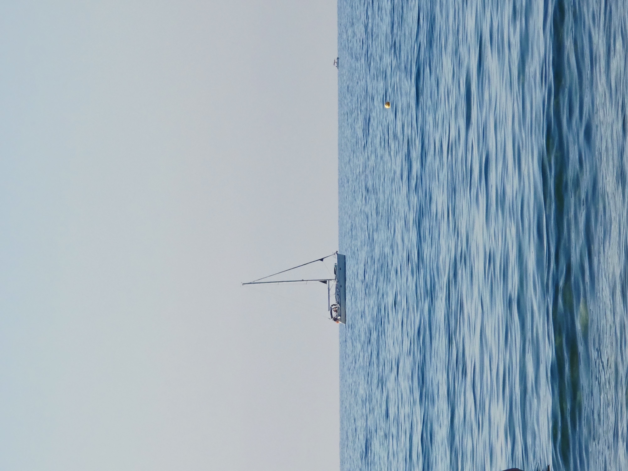 An image captured from the water shows a calm sea with gentle ripples. In the middle ground, a white sailboat with a single mast and dark sails is sailing horizontally. The boat has a visible cabin with people on deck. Further in the distance, a small white boat is visible on the horizon. A yellow buoy floats in the water between the sailboat and the distant boat. The sky is clear and a pale blue, indicating fair weather. The foreground of the image is dominated by the blue water, with the hint of land or a person on the far left. The scene is likely set in the waters off Malia Municipal Unit, Greece, a coastal area known for its beaches and sailing activities.
