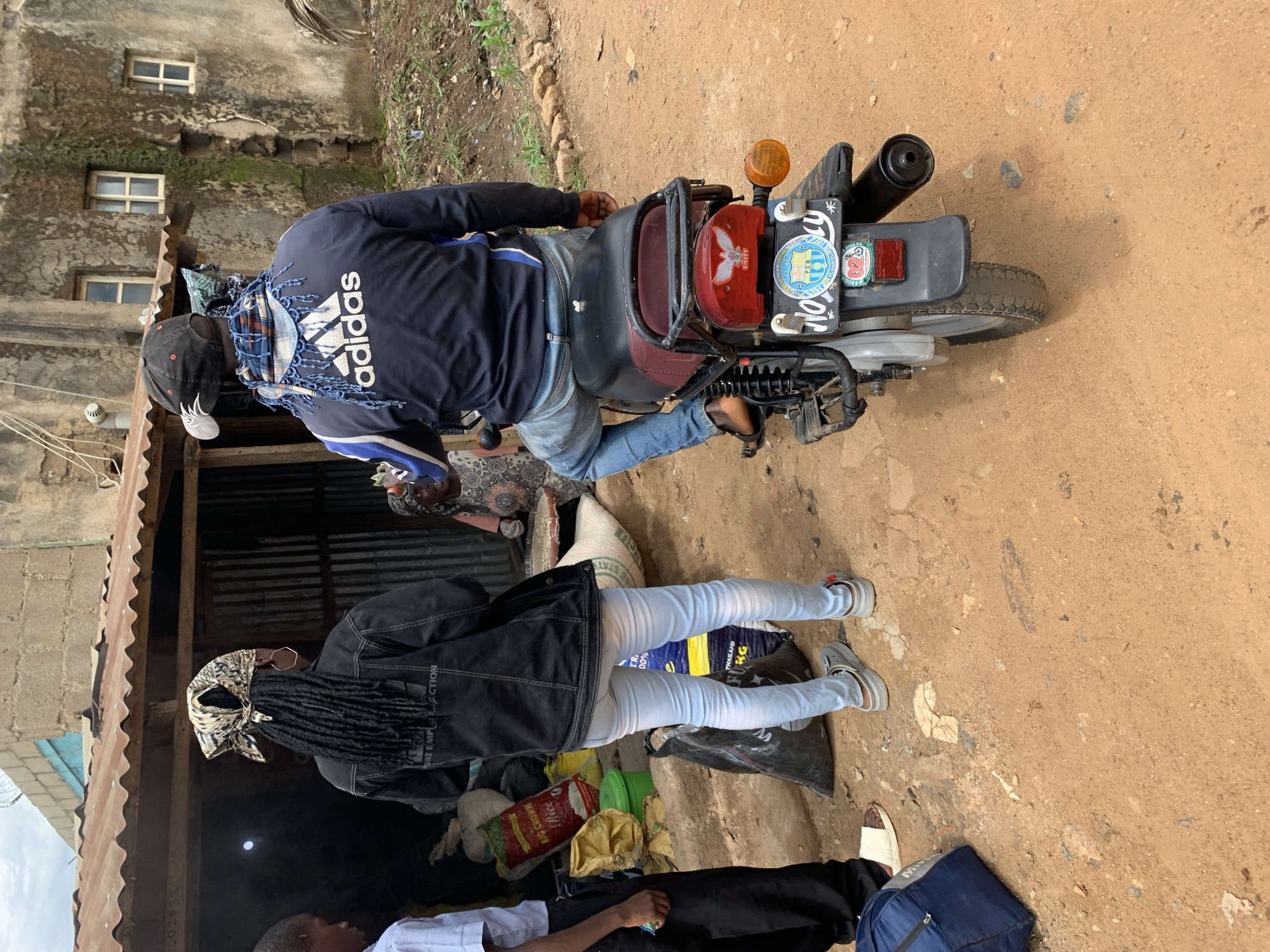 A ground-level image taken in Rafin Par, Nigeria, depicts three individuals and two structures on a dirt path under an overcast sky.
In the foreground-left, a young male stands wearing a light blue short-sleeved collared shirt and dark trousers. His right foot is bare, and his left foot is in a white slide sandal. A blue duffel bag rests on the dirt near his feet. In the mid-ground-center, a female is seen from behind, facing a stall. She has long dark dreadlocks gathered under a patterned headscarf (cream, brown, black). She wears a dark denim jacket with partial text "OWZ EMPL JACTION" visible on the back, and light blue skinny jeans. Her footwear consists of grey platform sandals with orange accents. In the mid-ground-right, a male is seated on a motorcycle, also viewed from behind. He wears a black baseball cap with a white design on the back, a blue and white plaid scarf draped over his shoulders, a dark blue Adidas track jacket with a prominent white logo on the back, and blue jeans. His feet, in dark sandals, rest on the motorcycle footpegs.
The female stands in front of a makeshift stall with a rusty corrugated metal roof, supported by wooden beams, and corrugated metal walls. Inside and around the stall, various sacks and bags are visible, including a red bag labeled "BIG CROWN Rice" and a large white sack. A black plastic bag rests on the ground near the female's feet. Behind the stall, a two-story building constructed from rough concrete or mud brick stands, showing signs of dilapidation with some exposed surfaces and moss/vines growing on its facade. It features multiple small, rectangular windows with white frames, some divided into panes. Barbed wire runs along the top of this building's wall on the left. The motorcycle has a black and red seat, a black rear luggage rack, and a red taillight assembly with a white bird/dove emblem above the word "UNITY". Below the taillight is a license plate featuring a round blue and white "FCB" (FC Barcelona) logo, and the alphanumeric sequence "W0400U" is partially visible. A black exhaust pipe extends from the right side, and an orange turn signal is visible.
The ground is unpaved, consisting of reddish-brown dirt and gravel, with scattered small stones and minor debris. The sky above is overcast and light grey.