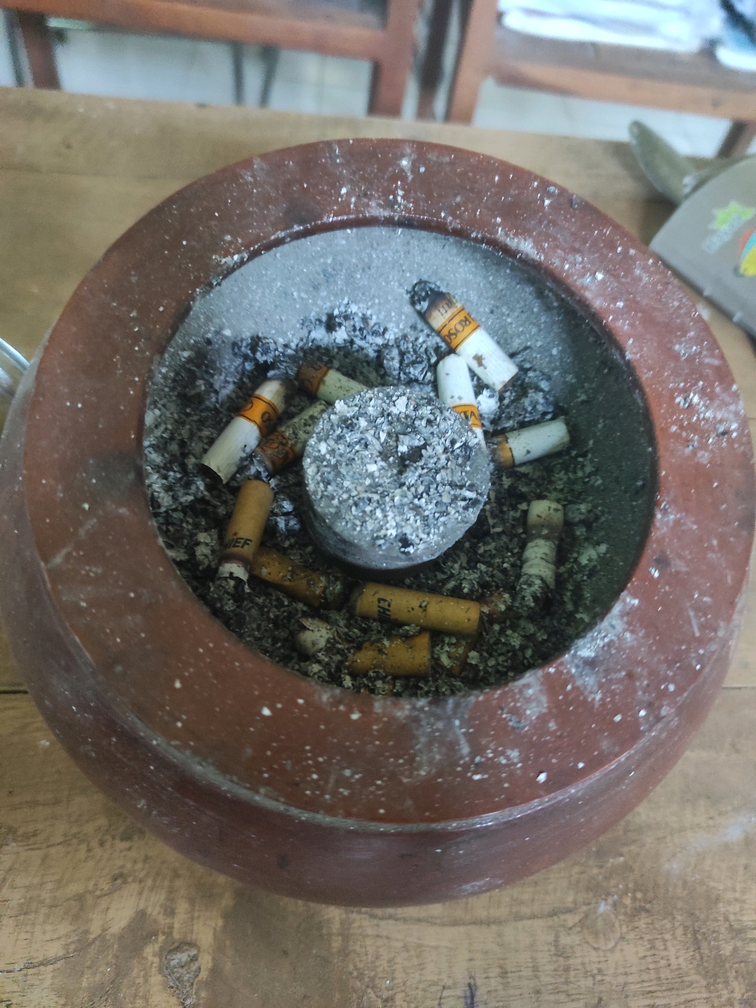 The image shows a close-up of an ashtray filled with cigarette butts and ash, placed on a wooden surface. The ashtray is made of a reddish-brown material, possibly clay or ceramic, and has a circular shape. Several used cigarette filters are visible, some with visible brand markings such as "ROSO" and "CHIEF". The surrounding wooden surface appears weathered and has a visible grain pattern. The lighting suggests it is indoors. The context of this scene in Ngujung, Indonesia, is not determinable from the image alone.