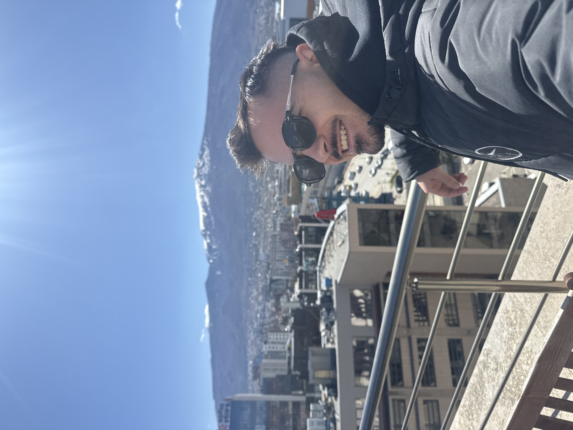 A man with a beard and sunglasses stands on a balcony overlooking Sofia, Bulgaria. He wears a black jacket with a hood. In the background, there is a cityscape with mountains and a blue sky. The balcony has metal railings.