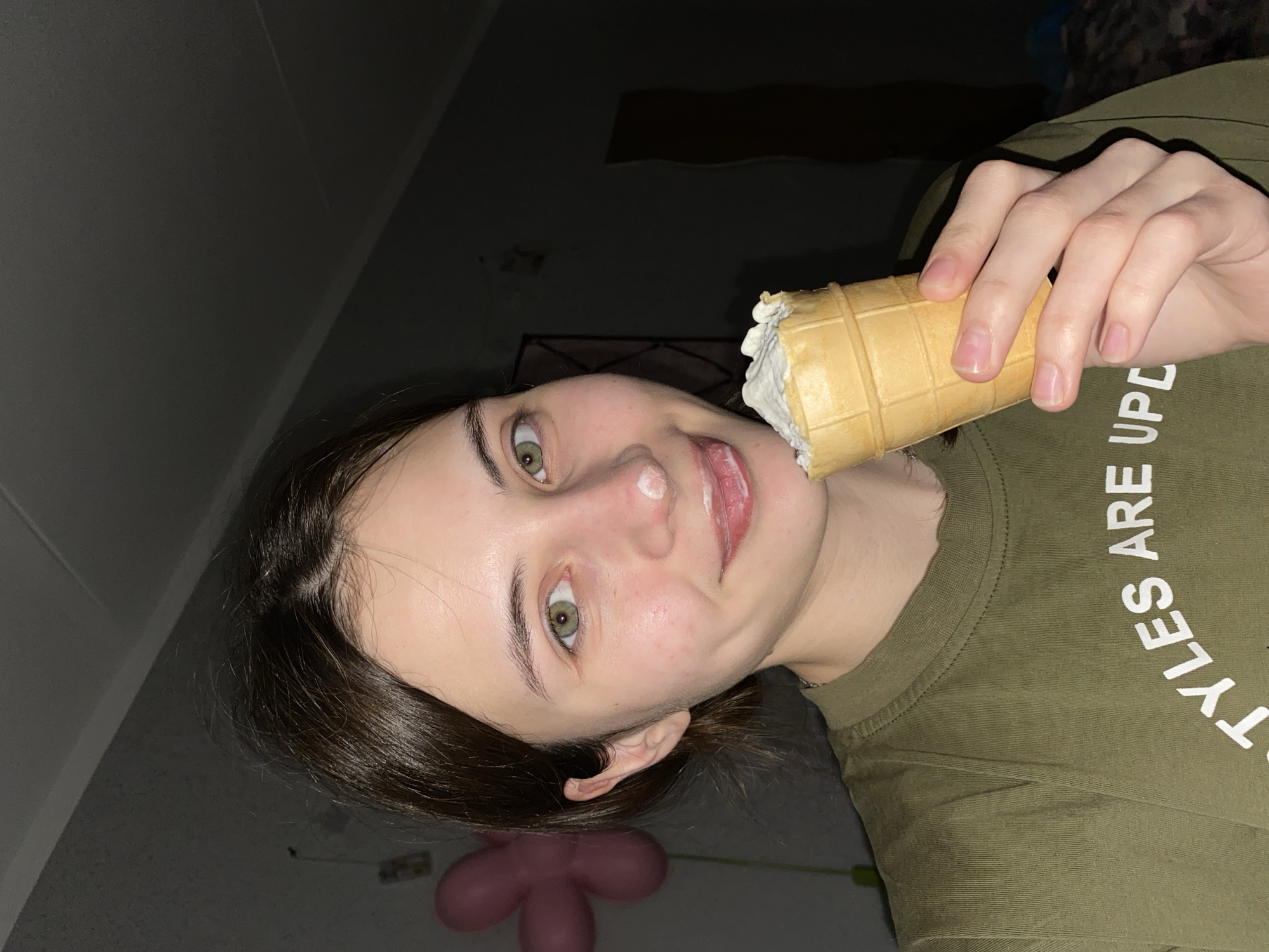 An eye-level indoor shot depicts a young person, likely female, with dark hair and striking green eyes, eating an ice cream cone. The subject is oriented at a tilted angle, as if looking up or lying down, with their head positioned towards the lower left of the frame. They are actively consuming a waffle cone filled with white ice cream, which has left a smudge on their upper lip and chin, indicating enjoyment. The person has a slight smile and their tongue is visible.
They are wearing an olive green t-shirt with partial white text visible on the right side, reading "...STYLES ARE UPD..." suggesting a phrase like "STYLES ARE UPDATED." The background is dark and indistinct, with hints of a ceiling or upper wall in the top left, implying a dimly lit indoor environment, possibly at night or in the evening. In the lower left corner, a soft, pink, multi-lobed object resembling a balloon is partially visible. No specific location cues for Votkinsk, Russia, are apparent in the image itself, which captures a moment of casual indulgence.