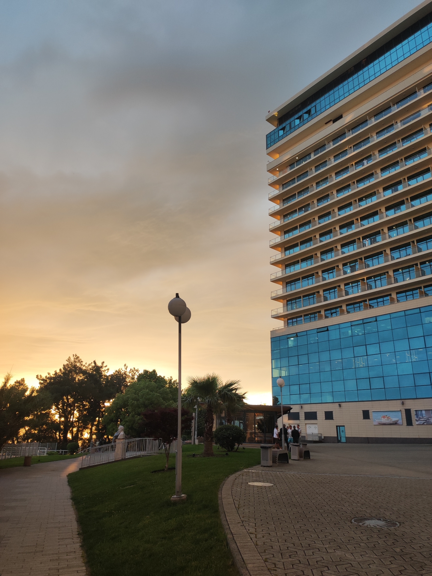The image depicts a view in the city of Sochi, Russia, likely near the Black Sea coast. On the right stands a tall, modern hotel with many floors, distinguished by its predominantly blue glass facade and balconies on each level. The lower section of the building features large panels of reflective blue glass, while the upper floors have repetitive balcony designs.
To the left of the hotel, the scene transitions into a landscaped area with trees and greenery. A pathway made of interlocking pavers curves into the distance, leading past a small garden with trees and a sculpture atop a low bridge.
A street lamp with two globes is positioned in the foreground. Closer to the hotel, there is a paved area with several people grouped together, possibly engaged in conversation.
The sky is filled with an orange and grey hue, suggestive of sunset or sunrise. The overall impression is one of a well-maintained public space juxtaposed against the modern architecture of the hotel, set against a scenic sky.