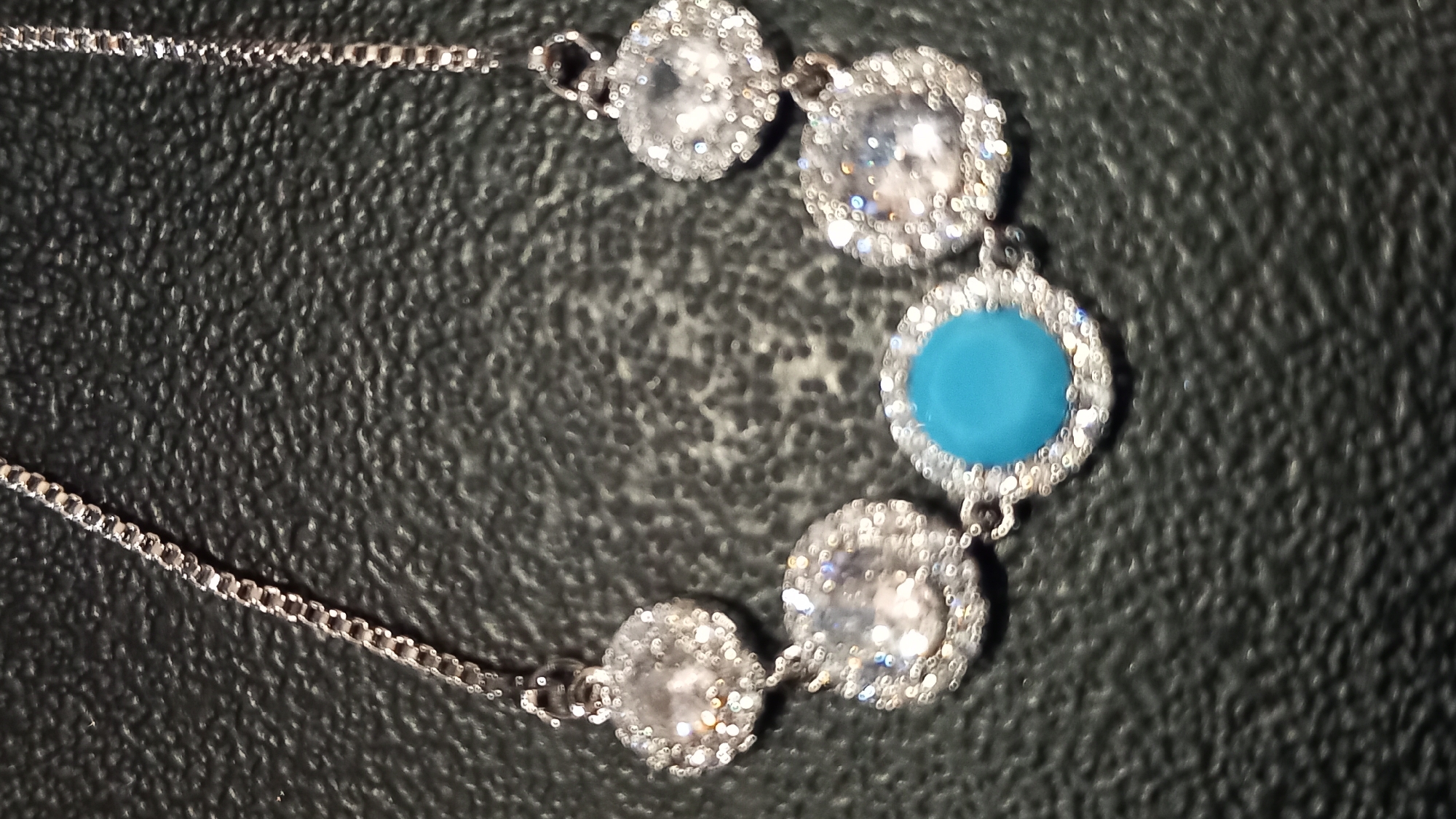 The image shows a close-up, top-down view of a silver necklace with several clear, round stones and a single, round, turquoise stone. The necklace is displayed against a dark, textured surface that appears to be black leather or a similar material. The chain is made of small, interlocking links. The clear stones are set in circular bezels that are encrusted with smaller, glittering stones, likely diamonds or cubic zirconia. The turquoise stone is also set in a similar bezel. The lighting is bright, causing reflections and highlights on the stones and chain. The image quality is slightly blurry due to the focus. There are no people, animals, or any other objects visible. The image does not provide any information about its location within the city of Kos, Greece.
