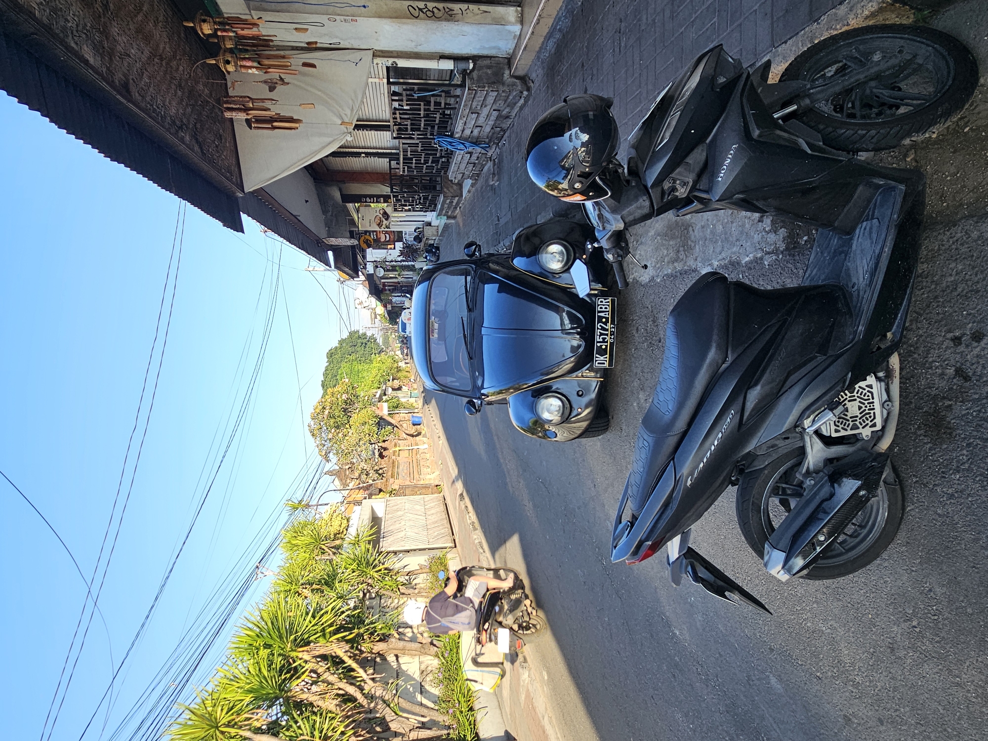 A street in Denpasar, Indonesia, is captured under a clear blue sky. The scene is dominated by two parked motorcycles in the foreground and a black Volkswagen Beetle in the middle ground. The motorcycle closest to the viewer is black, with a black seat and visible "HONDA" branding on its front panel. The second motorcycle, a scooter, is further back on the left, ridden by a person wearing a white helmet, dark t-shirt, and shorts, carrying a light-colored bag on its rear rack.
The Volkswagen Beetle, a classic model, is parked on the right side of the road, displaying a license plate starting with "DK" followed by numbers and letters. It's positioned on a paved road, with a sidewalk running alongside it. The sidewalk is lined with shops and businesses, some with corrugated metal roofs. Wind chimes made of bamboo are hanging from one of the shop awnings.
Vegetation, including palm trees and leafy shrubs, borders the left side of the street, casting shadows on the road. The buildings on the right appear to be single-story structures with shopfronts. The overall impression is a typical street scene in a tropical urban environment, likely during daytime given the bright sunlight and clear sky.