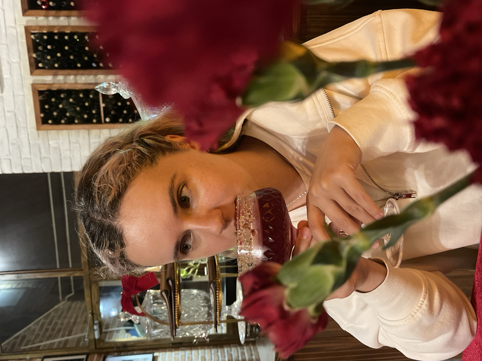 A person is seen holding a glass of red liquid, possibly a beverage, close to their face. They are wearing a light-colored zip-up jacket. The foreground is partially obscured by red flowers, likely carnations.
In the background, a wall with framed displays is visible, along with an interior space featuring decorative items and possibly a bar area. A reflective surface suggests a mirror or glass display case. The overall lighting suggests an indoor setting, likely a restaurant or bar.
Without additional information, specifically elements definitively linking to Lâm Viên - Đà Lạt, it's impossible to confirm this location based on the visual elements alone.