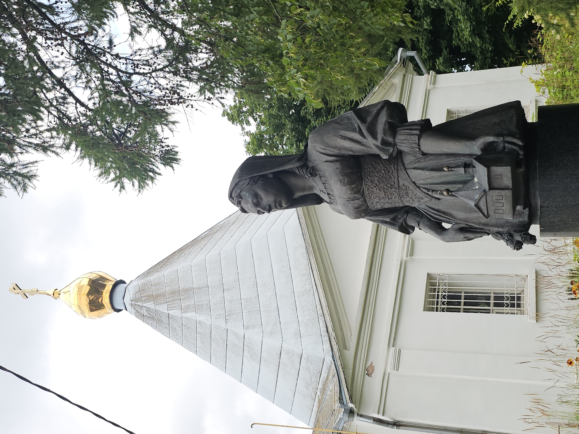 The image shows a bronze statue of a woman holding a miniature building in front of a white church with a golden dome. The statue is of a woman dressed in historical clothing, with her head covered by a veil and her hair styled in curls. She is wearing a long, flowing dress with intricate detailing on the bodice and sleeves. In her hands, she holds a small model of a church, complete with a dome and windows. The church in the background is a white, rectangular building with a stepped roof and a golden dome topped with a cross. A small, barred window is visible on the side of the church. The scene is set outdoors, with lush green trees and foliage surrounding the church and statue. The sky is overcast with a few wisps of clouds. This statue and church are located in the village of Ulitkino, Russia, a suburb of Moscow.