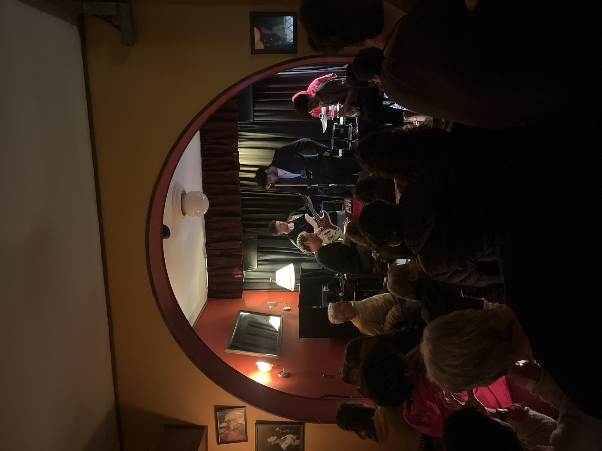 A photograph depicts a dimly lit interior space, viewed through an arched opening. The image appears to be rotated or taken at an angle. The space features a stage where a musical performance is taking place. Musicians are visible, including a drummer, a bassist, and a guitarist. A small audience is present, gathered closely to watch the band. The walls are painted in dark colors, adorned with framed pictures and sconces. A large white globe-shaped lamp hangs from the ceiling. The scene is reminiscent of a small, intimate venue. It is highly possible that this place is located in the city of Buenos Aires, Argentina because the architecture and the paintings on the wall are Argentinian.