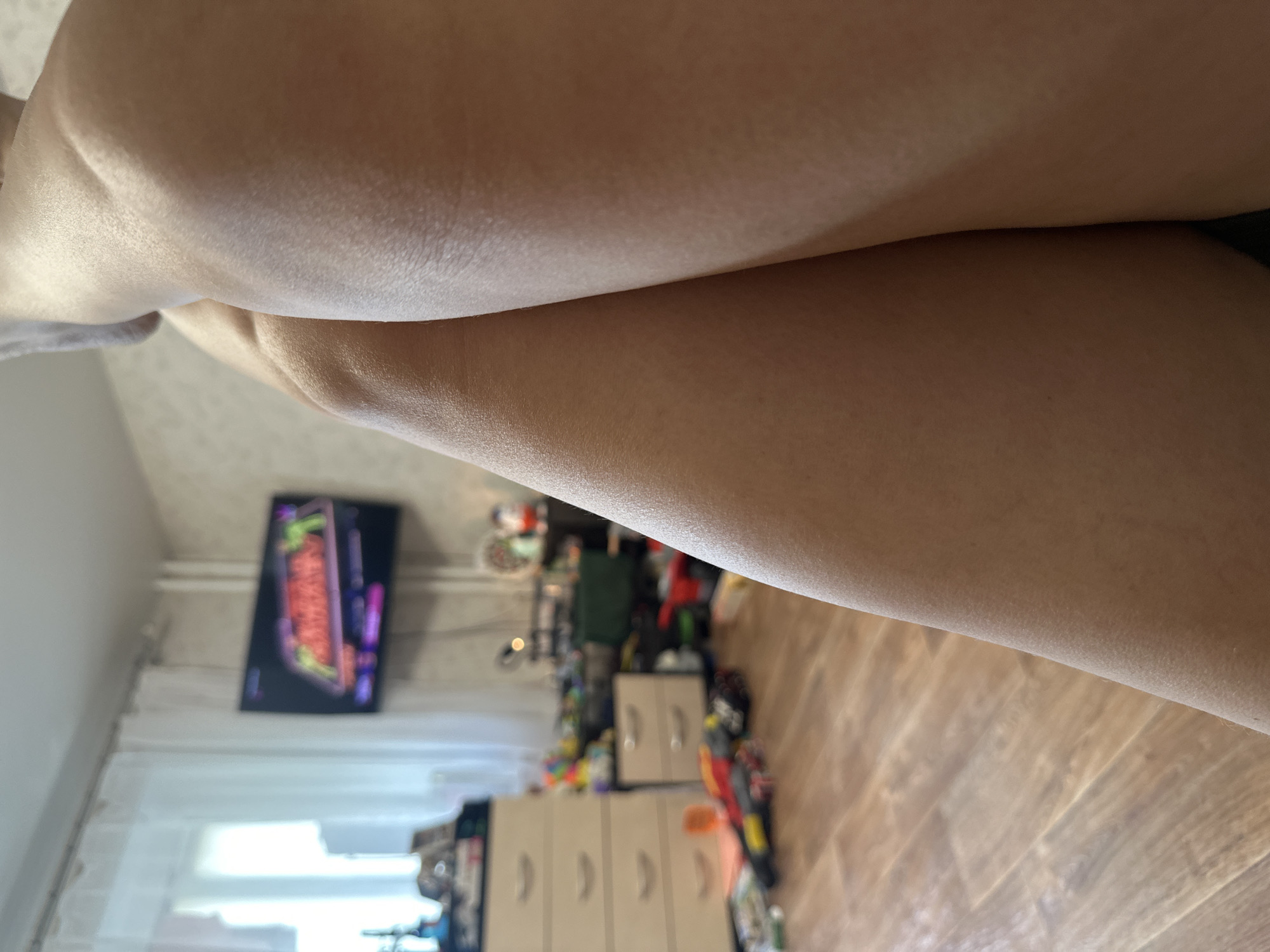 The image shows a close-up view of a person's legs, with the lower body and thighs prominently featured. The skin appears smooth and fair. The background is blurred, but suggests an indoor setting, possibly a room in a house. There are hints of furniture and possibly a television screen on the left side of the image. The lighting suggests it could be daytime, with natural light entering the room. The overall impression is a casual and personal snapshot. The image is oriented sideways.