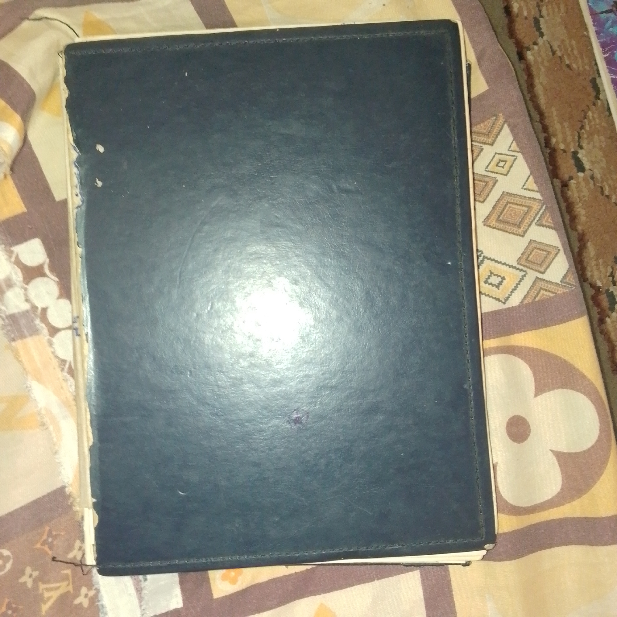 The image shows a closed, dark blue binder or book lying on a patterned fabric surface. The binder has a textured cover, possibly faux leather, with visible stitching along its edges. There is a bright reflection on the binder's surface, suggesting a light source from above and to the left. The fabric beneath has a repeating pattern of brown, yellow, and beige shapes, including diamond-like forms and abstract floral elements. A small, dark purple stain is visible on the binder's cover. The binder appears to be aged, with some wear and tear on its edges. The overall scene is presented from a top-down perspective. The image was captured in Jalingo, Nigeria.