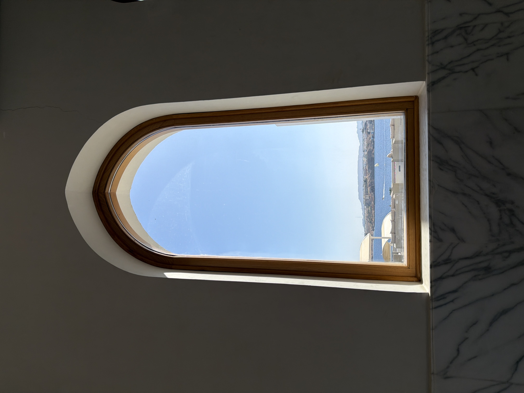 A vertically oriented window with a gently arched top and a straight bottom provides a bright view of the outdoors from an interior setting. The window features a warm, light brown wooden frame. The wall immediately surrounding the window is a plain, light off-white or beige color, showing subtle hairline cracks in the upper left section. To the right of the window, a vertical section of the wall is covered in white marble with prominent dark grey veining.
Through the clear window glass, a vibrant blue sky fills the upper portion of the view, transitioning to a deep blue body of water, likely the Red Sea, in the middle and lower sections. Along the distant horizon, a strip of land with various buildings is visible, suggesting a coastal development. Scattered white structures, possibly parts of buildings, balconies, or boats, can be seen near the water. The weather is bright and sunny, indicative of midday or early afternoon, with strong natural light illuminating the exterior scene and contrasting with the shadowed interior wall. Some minor smudges or reflections are visible on the window glass.
No people or visible text are present in the image. The scene offers a tranquil glimpse of the landscape, characteristic of a coastal resort area such as Sharm El Sheikh, Egypt.