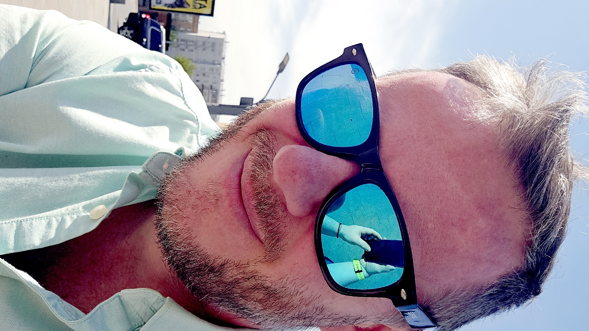 A medium, angled close-up shot depicts a fair-skinned man with short gray hair, stubble, and a button-down mint green shirt. He is wearing black sunglasses with blue reflective lenses. The reflection in the sunglasses appears to show a hand holding a black smartphone, with a lime green wristband on the arm. The background, visible behind the man, includes city elements such as buildings, a street lamp, a vehicle, and a sign. The angle of the shot suggests the man is looking up. The location is possibly Moscow, Russia, given the architectural style of the buildings and signage.