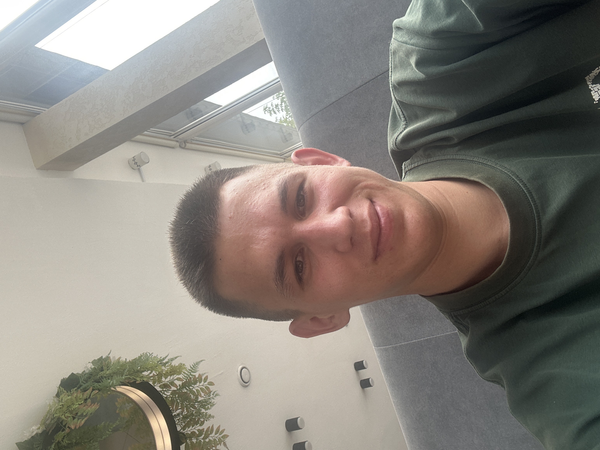 A young Caucasian male with short brown hair and fair skin is captured in a selfie taken from a slightly high angle. He is wearing a dark green t-shirt. The background suggests an indoor setting with modern architecture. To the left, a hanging plant with green foliage is partially visible against a white ceiling with recessed lighting fixtures and what appear to be black cylinder-shaped lights. To the right, a large window with white framing reveals a bright, overcast sky and the suggestion of trees outside. A light-colored, textured structural beam divides the window, and a section of a gray upholstered bench or seating area is visible behind the man. The overall environment appears to be a cafe or public space in Oral, Kazakhstan, characterized by contemporary design elements.