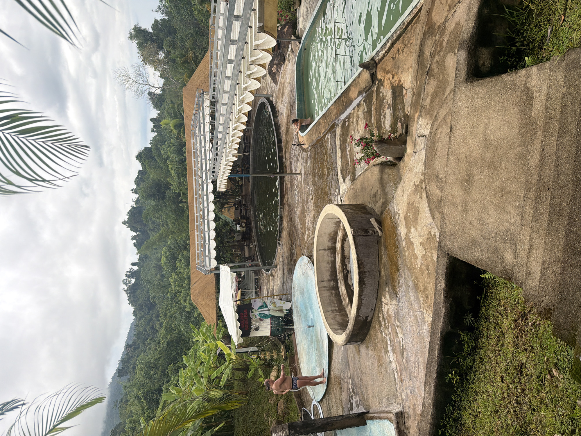 In Khanh Vinh, Vietnam, an outdoor recreational area features several pools. A man in swimming trunks stands by a shallow, circular pool with a fountain in its center, holding a hose. Adjacent to this is a larger, oval-shaped swimming pool filled with green water. Further in the background, a large, dark circular pool is visible beneath a covered walkway. The landscape is lush, with dense green foliage and trees covering the hillsides surrounding the area. The sky is overcast.
