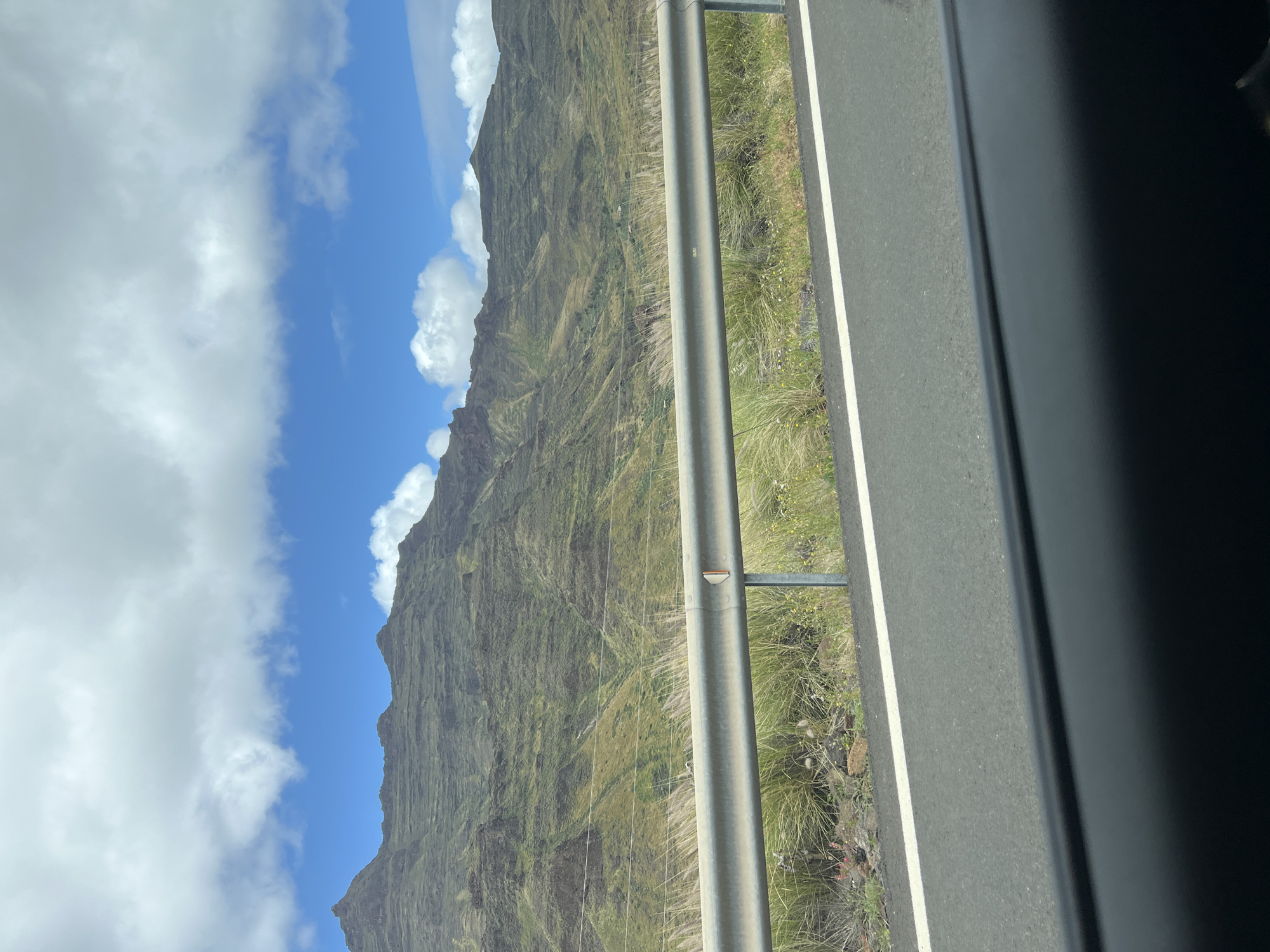 A view from a vehicle traveling on a roadway in Mogán, Spain. The window frame of the vehicle is visible on the right side of the image. A guardrail runs parallel to the roadway, separating the road from a steep mountain slope covered in green vegetation. The sky is blue with patches of white clouds. Power lines are visible against the mountain slope. The road surface is gray with a white dividing line.