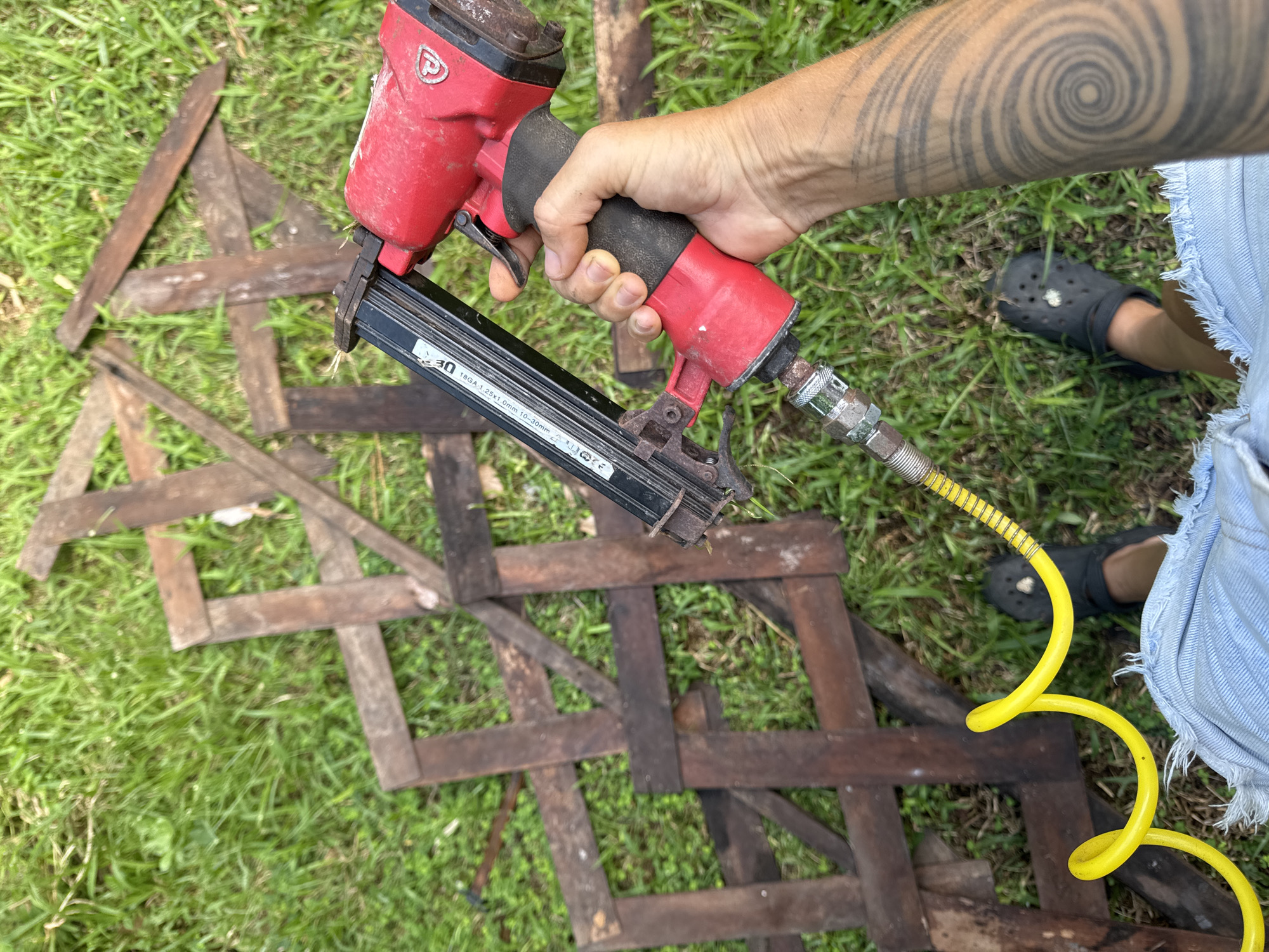 The image presents a close-up, high-angle view of a person holding a red pneumatic nail gun outdoors in Kediri, Indonesia. The person's right hand firmly grips the tool, which is connected to a bright yellow coiled air hose. The arm above the hand features a large, striking spiral tattoo.
The nail gun is pointed downwards towards a collection of dark, weathered wooden slats scattered across a vibrant green grassy ground. Some of these wooden pieces form an informal, crisscrossed pattern, suggesting either materials for a project or discarded elements. The person's lower left leg, clad in light blue ripped denim shorts and a black Croc shoe, is visible towards the bottom right, further indicating an outdoor, casual work setting.
Visible text on the side of the nail gun's magazine specifies its type and capacity: "18GA 1.25x1.0mm 10-30mm", indicating it's an 18-gauge nailer capable of firing nails between 10mm and 30mm. A brand logo, appearing as a stylized "P" in a circle, is on the main body of the tool. The bright and clear lighting suggests it is daytime with good weather conditions. The scene captures a moment during what appears to be a carpentry or construction activity.