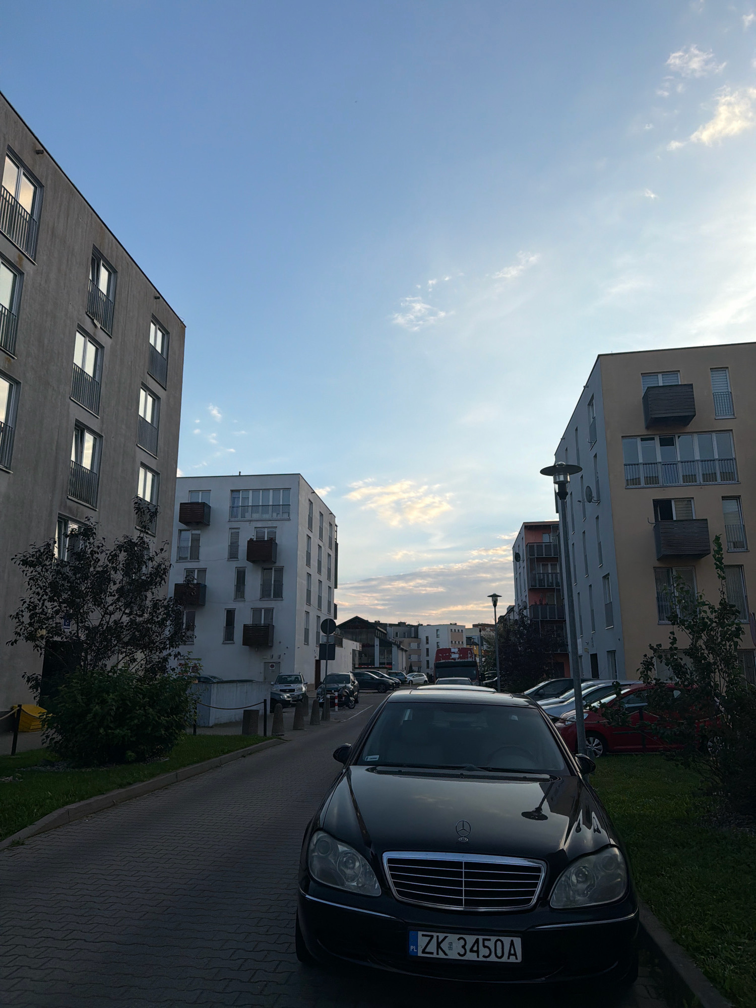 The image shows a street in the city of Koszalin, Poland, lined with modern apartment buildings. A black Mercedes-Benz S-Class sedan with license plate ZK 3450A is parked on the right side of the street, facing the viewer. The car is detailed, showing its chrome grille, headlights, and the Mercedes-Benz emblem. To the left of the car, a row of cobblestones forms the pavement, leading up to a green lawn and a dark, leafy tree.
The buildings are multi-story with balconies and numerous windows. The building on the left is gray, while the building in the center is white, and the building on the right is a light yellowish-beige. The sky above is a clear blue with scattered white clouds, suggesting it is either early morning or late afternoon. Streetlights and some other vehicles are visible further down the street. The overall scene depicts a residential area in an urban setting.