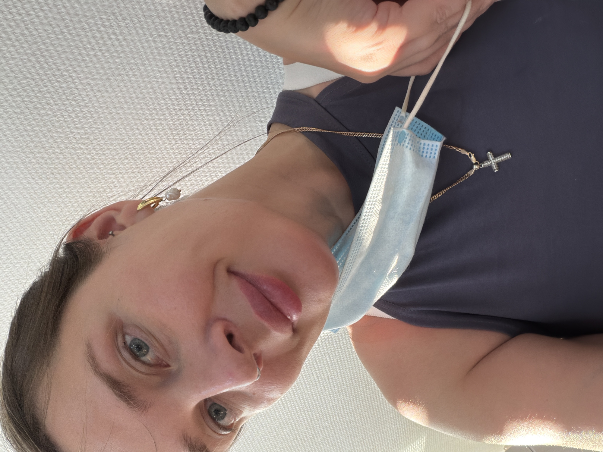 The image is a close-up, low-angle portrait of a person, likely a woman, looking directly into the camera with a soft, relaxed expression. Her head is tilted slightly, and her light-colored eyes (blue/grey) are focused downwards.
She is wearing a dark blue or grey top. A light blue surgical mask hangs below her chin, suspended by its ear loops from a gold chain necklace. This necklace also features a silver-toned cross pendant adorned with small, sparkling stones. Another delicate gold chain necklace with a small, light-colored pendant, possibly a pearl, is visible around her neck. In her left ear, she wears an earring with a gold element and a white pearl drop, and she has a small nose ring (likely a septum ring). On her left wrist, a black beaded bracelet is visible. A hand is partially visible at the top of the frame, holding the mask's ear loops.
The lighting is bright, suggesting daytime conditions, and it illuminates her face and the upper part of her body. The background is a uniformly bright, off-white surface with a subtle, textured pattern, possibly a ceiling or a plain wall, indicating an indoor setting. No text is visible in the image.