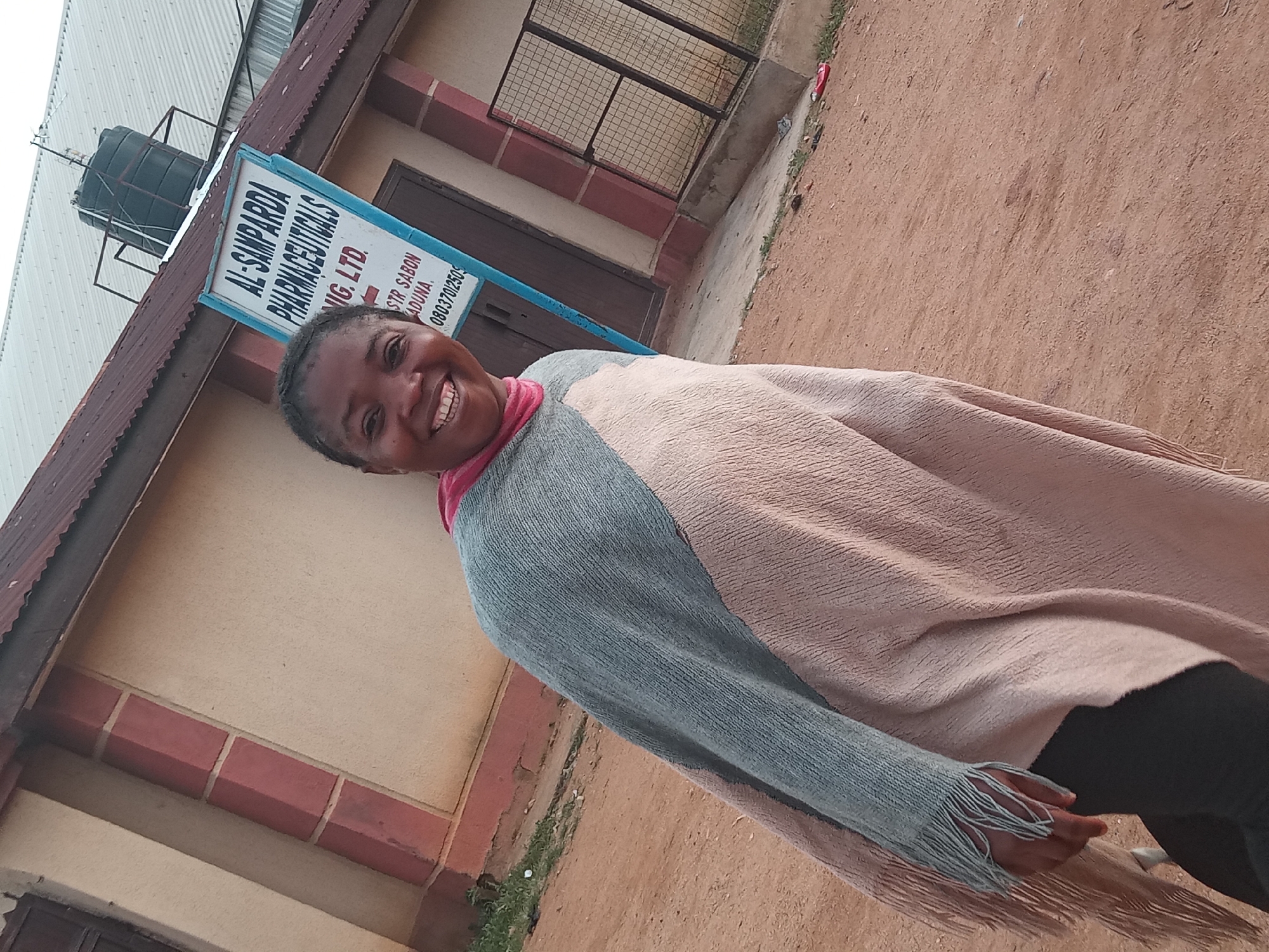 A medium shot captures a Black woman from the shoulders down, smiling and looking at the camera, with a building and a sign in the background. The woman is wearing a pink scarf and a large, colorful poncho made of woven fabric with fringe. The poncho is predominantly light pink and light blue. Her left hand is visible, resting on her hip. The building behind her is light brown with reddish-brown accents and a corrugated metal roof. A blue and white sign with text in black lettering is mounted on the side of the building. The text on the sign appears to be "AL-SAMPARDA PHARMACEUTICALS NIG. LTD." and below that "STR. SABON TADUNA." followed by a phone number. The ground in front of the building is dirt and gravel. The image is taken outdoors, likely in the city of Sabon Tasha, Nigeria, given the signage.