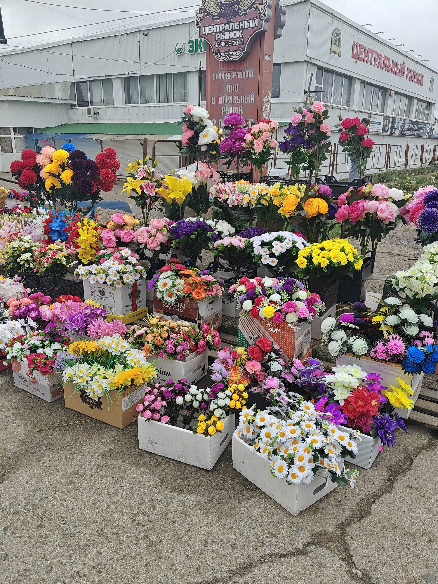 A vibrant array of artificial flowers is displayed in boxes and stands in front of the "Central Market" in Chaikovsky, Russia. The flowers include roses, lilies, daisies, and other varieties in a wide range of colors. The market building is visible in the background, with signage in Russian. The street is paved and appears wet in some areas.