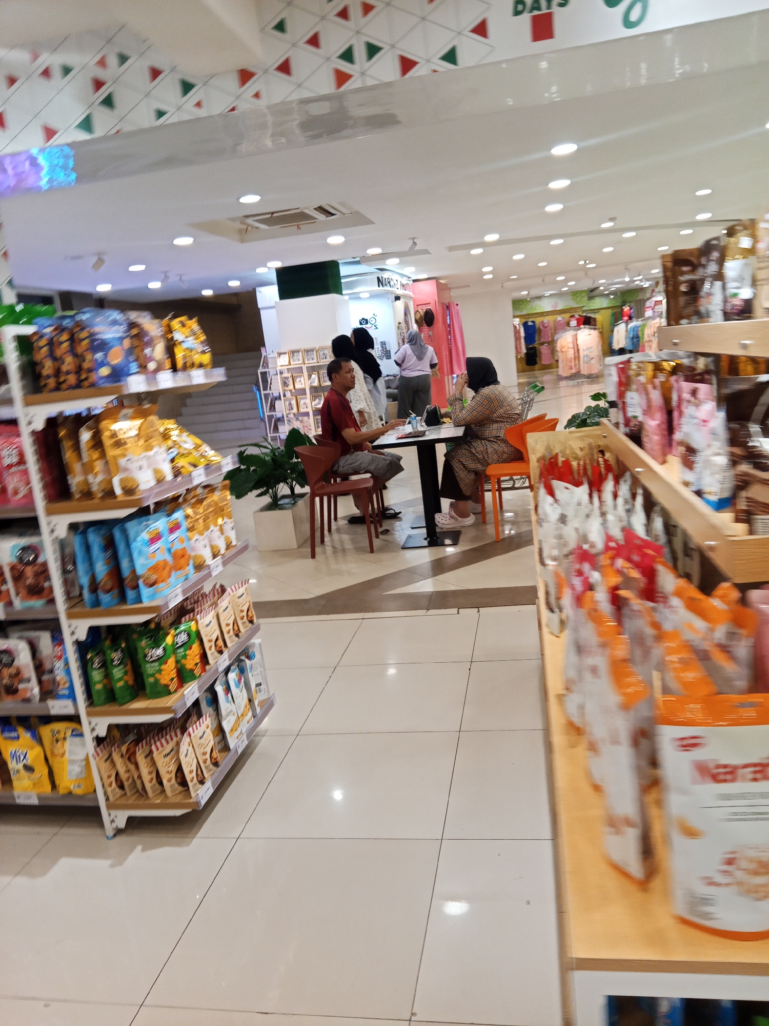 The image depicts the interior of a supermarket in Lubuklinggau, Indonesia, possibly in the Citimall. Aisle shelves filled with packaged snacks and food items are visible on the left and right sides of the frame. In the mid-ground, two individuals are seated at a table, appearing to be engaged in a conversation or a break. One person is wearing a plaid patterned outfit and a hijab, while the other is wearing a red shirt and gray pants. A third person, wearing a pink top and gray pants, is standing near the table. The environment is well-lit with overhead fluorescent lights and shows typical retail store features like flooring and ceiling design.