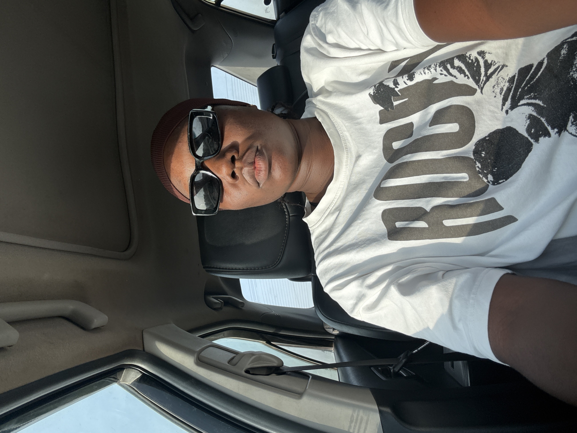 A dark-skinned person wearing a brown beanie and black sunglasses is sitting in a car. The person is wearing a white t-shirt with black writing and a black and white graphic. The car's interior is gray, and a seatbelt is visible. The image suggests the car is parked or stationary. There's no information about Kuchingoro, Nigeria.