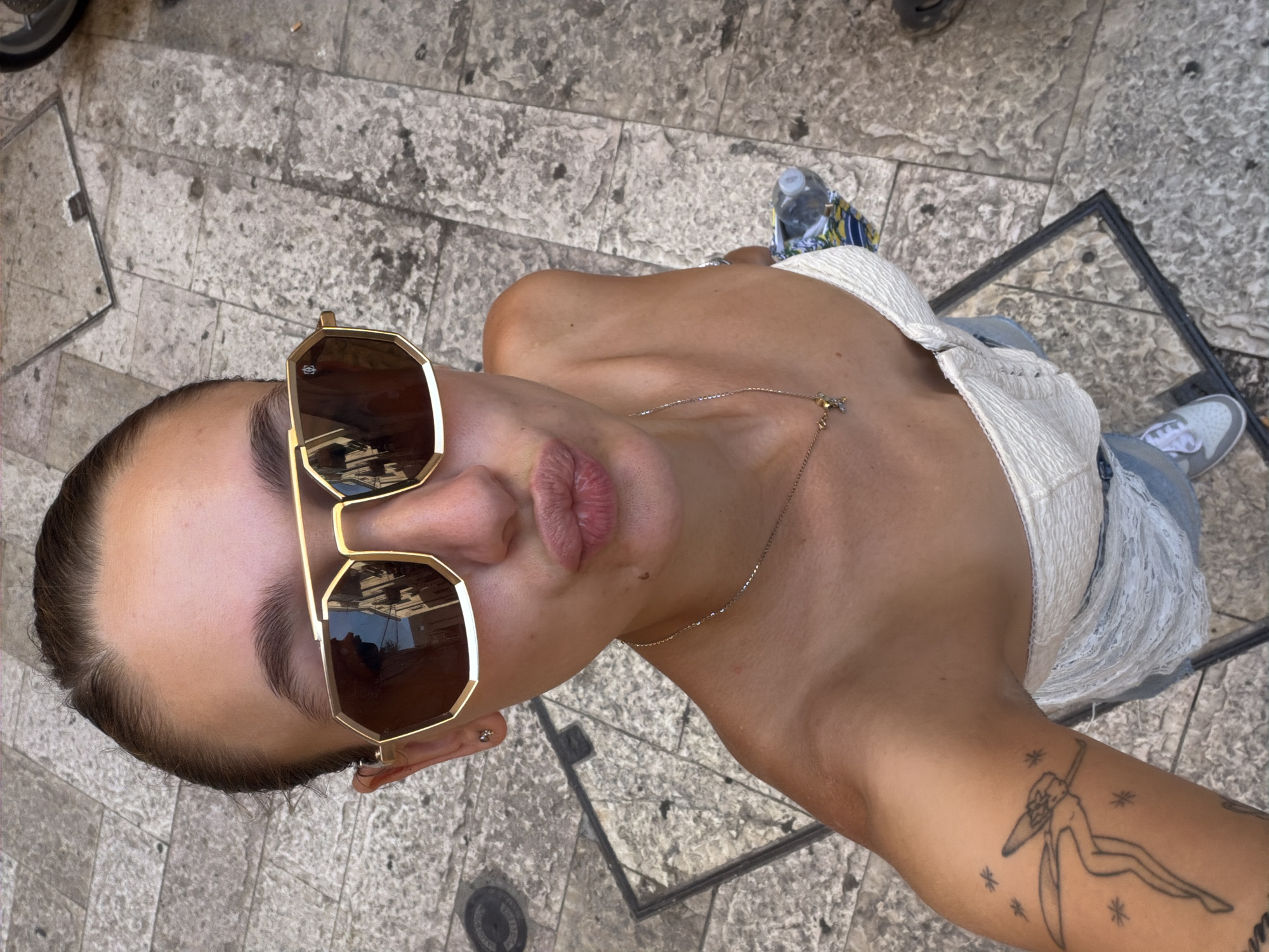 In Bari, Italy, a young woman is captured in a high-angle selfie taken outdoors. She is wearing large, gold-rimmed sunglasses and a strapless white top. Her lips are puckered as if blowing a kiss. She has a delicate necklace with a small pendant. On her left arm, a tattoo of a fairy with outstretched arms and small star-like shapes is visible. Her hair is pulled back. The ground beneath her is paved with large, light-colored stones, characteristic of many Italian cityscapes, and a metal grate is visible in the foreground. A partially visible water bottle and a patterned fabric are on the ground to her right. The overall setting suggests a sunny day in a pedestrian area of the city.