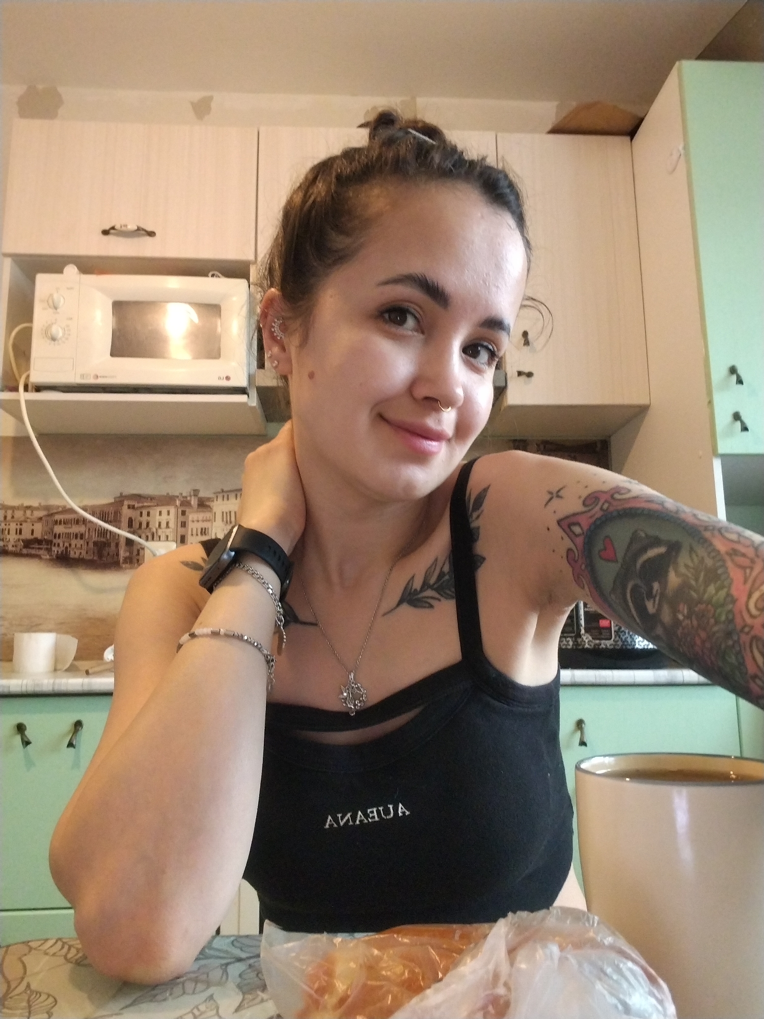 A woman is indoors in a kitchen setting. The kitchen has light-colored cabinets with dark handles. A microwave is positioned above a cityscape mural. The woman has her hair in a bun, with visible tattoos on her left arm and collarbone. She is wearing a black tank top and jewelry including a necklace, bracelets, and ear piercings. A mug and a plastic bag containing food are visible on the table in front of her.
The city of Noviy, Russia, is not visible in the image. The cityscape mural may be of another city.