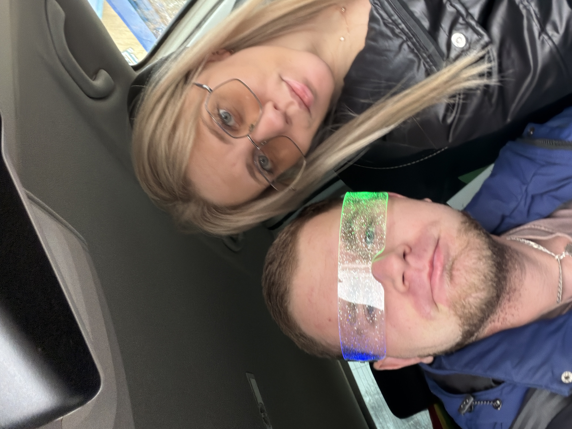 Two people, a blonde woman with glasses and a man with a beard, are seated in a vehicle. The woman is wearing a dark jacket, and the man is wearing a blue jacket with a silver chain around his neck. A light artifact is present on the man's face. The interior of the vehicle is visible, including the dashboard and window. The location is presumably in a city, based on the suggestion of "Chaikovsky, Russia" in the request.