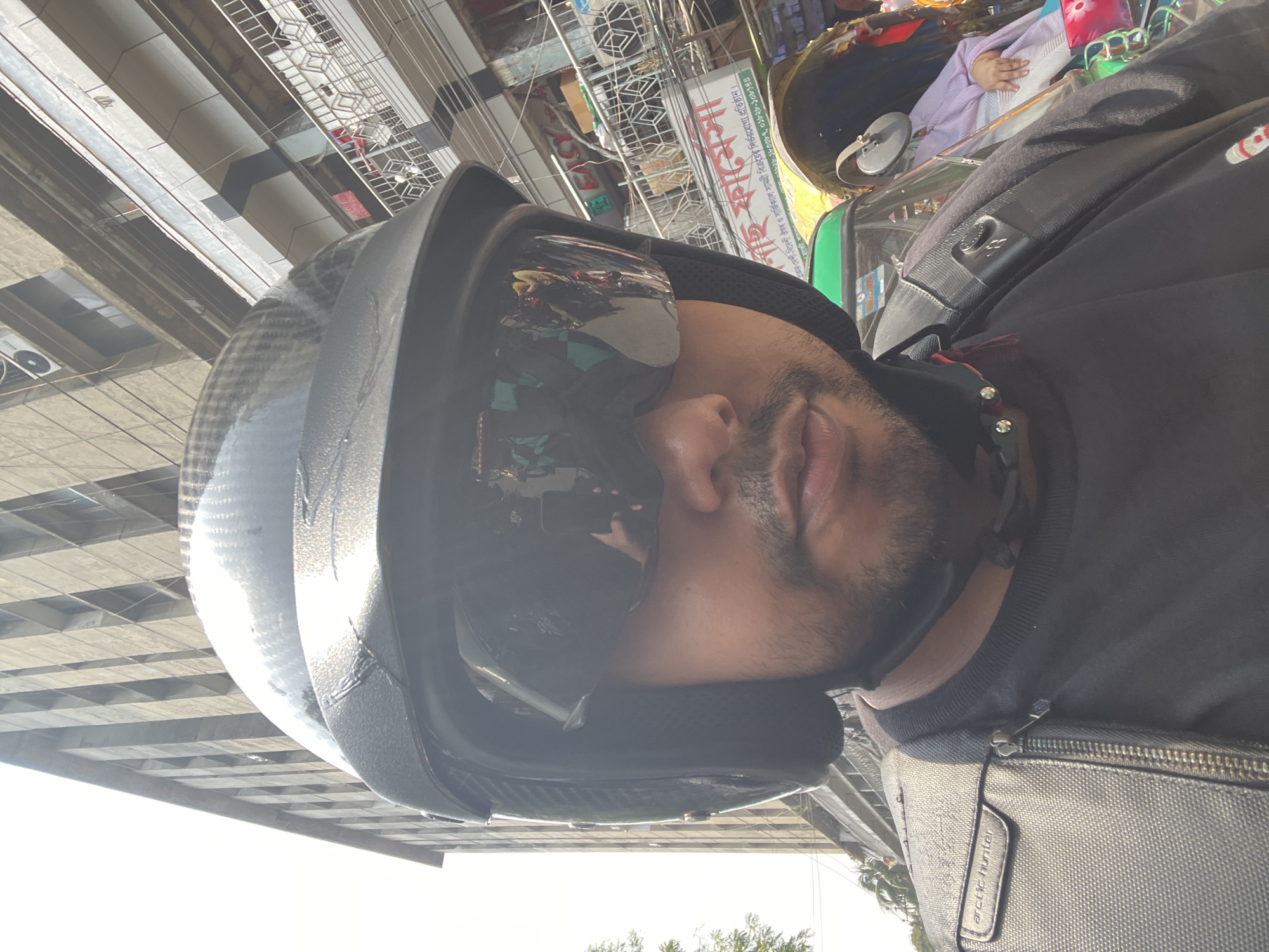 A close-up shot presents a person wearing a black motorcycle helmet with a reflective visor. The reflection on the visor obscures the facial details but reveals elements of the surrounding environment. The person is also wearing a black jacket or sweatshirt and a gray shoulder bag labeled "arctic hunter".
The background indicates an urban setting. Buildings, possibly apartment complexes or commercial structures, are visible. A signboard in the background contains text in Bengali and English, advertising medical supplies and pharmaceutical products. Part of the sign reads "নাই ফামেসা প্রকার দেশী-বিদেশী ঔষধ ও সার্জিক্যাল সামগ্রী বিক্রয়ের নির্ভরযোগ্য প্রতিষ্ঠান", which translates to "Nai Famesa - A reliable establishment for selling various local and foreign medicines and surgical supplies".
Based on the Bengali script on the sign, the location is most likely in Dhaka, Bangladesh. The composition of the shot suggests it might have been taken as a selfie or from a similar first-person perspective.