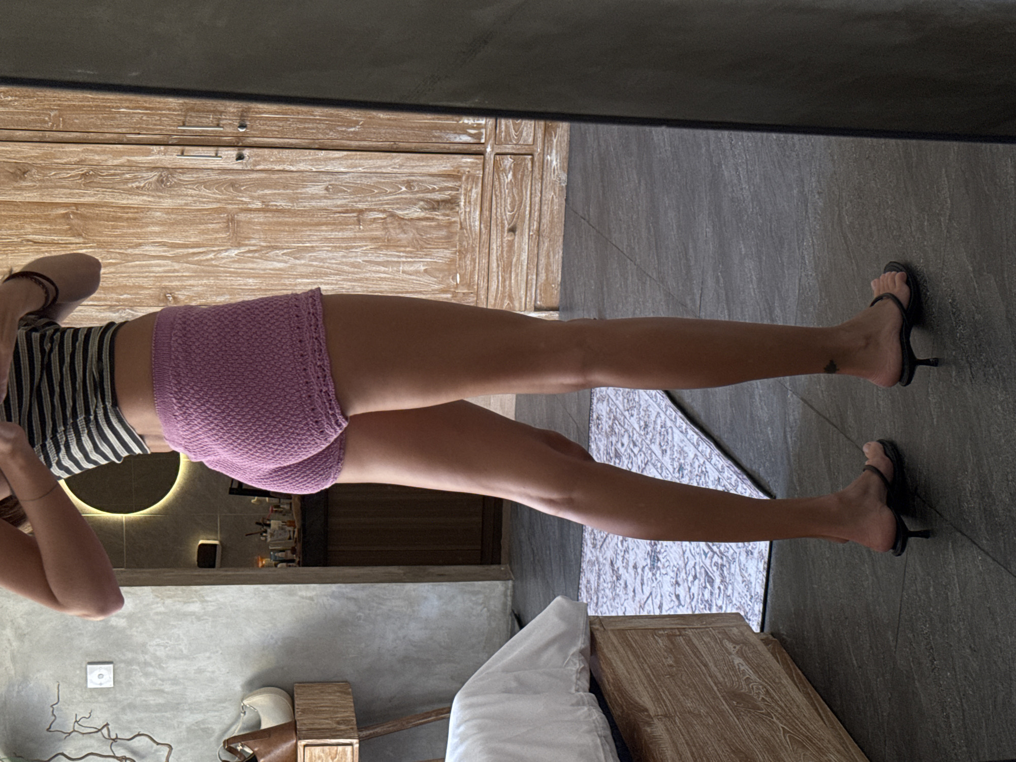 A woman wearing a striped tank top and pink knit shorts stands in a room with a light wood dresser. She is wearing black heeled sandals. The room has a grey tile floor and a grey rug with a geometric pattern. A bed with white sheets is in the lower right corner of the frame. A light-up vanity mirror and toiletries are visible in the background, which suggests this is a hotel room in the area of North Kuta, Indonesia.