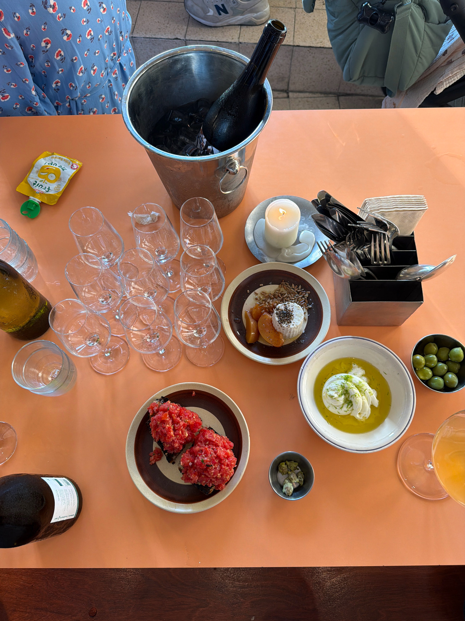 This is an overhead shot of a table with food and drinks, likely at an outdoor restaurant in Tel Aviv, Israel. The table is covered with an orange tablecloth. On the left, a bottle of dark wine is in an ice bucket with ice and a black cloth. Next to it, several empty wine glasses are arranged. A small yellow pouch with the word "fruit" on it is also visible.
In the center of the table, there are several plates of food. One plate has two servings of what appears to be a tomato and red pepper dish on toast. Another plate contains a soft white cheese with grains and what looks like dried apricots. A small bowl of olives and a larger bowl with a creamy dish topped with herbs are also present. A candle is lit on a small plate, and a set of cutlery is in a holder.
On the right side of the table, a glass of an orange beverage is visible, along with some other small dishes. In the background, a person wearing a blue dress with a floral pattern is partially visible, as is a light green jacket or bag and a pair of grey sneakers. The overall setting suggests a casual dining experience in Tel Aviv.