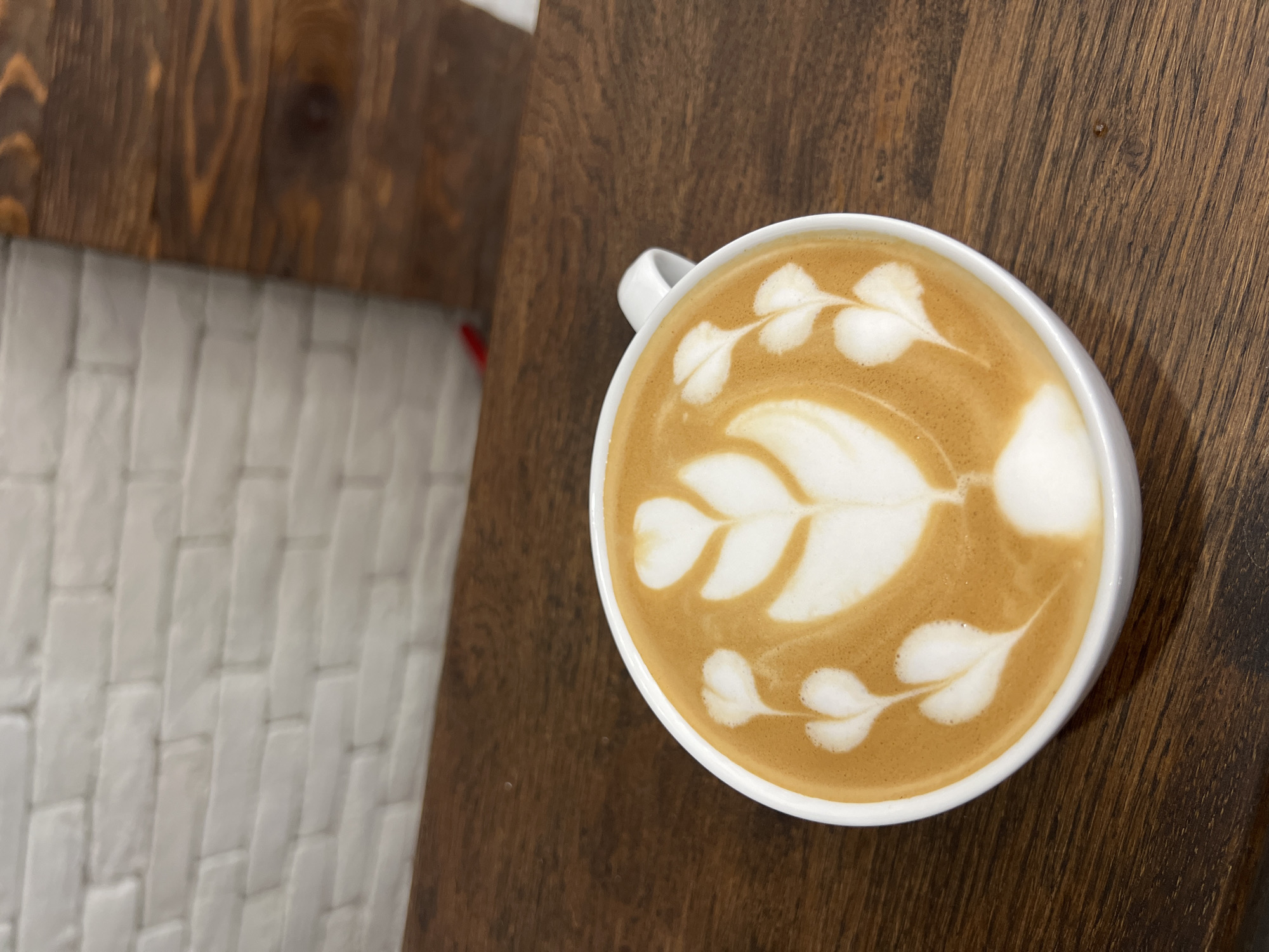 A high-angle shot shows a white ceramic mug of latte art, set on a dark brown wooden surface. The latte art design features a central leaf shape with additional leaves arranged around it. The background includes a partial view of a white brick-like wall on the left, and what appears to be another section of the wooden surface. The city and precise location are unknown.