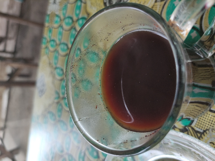 A close-up, top-down shot features a clear glass mug holding a dark brown liquid, likely coffee. The liquid shows a subtle reddish-brown gradient towards the bottom and edges. Visible coffee grounds or residue cling to the inner walls of the glass, particularly above the liquid line, indicating a fresh brew or a beverage where sediments are common.

The mug is placed on a distinctively patterned surface, which appears to be a tablecloth or mat. This surface exhibits a repetitive motif of light green, oval, or eye-shaped designs, outlined in a darker shade, set against a background of yellow and brown hues. Some sections of the pattern include smaller dots or textural details.

The background is softly blurred and out of focus, showing indistinct shapes and muted colors. This suggests an indoor or shaded outdoor setting. No individuals or specific activities are depicted; the image is a static capture of the beverage. The lighting appears natural, consistent with daytime. There is no visible text within the frame. The image is captured with a horizontal orientation, effectively rotating the cup 90 degrees.