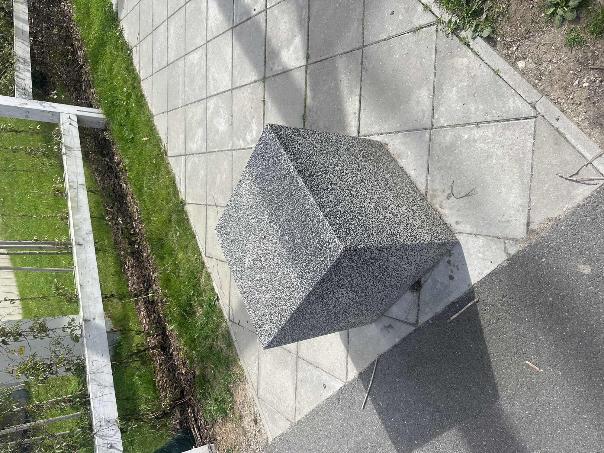 A medium shot captures a grey speckled cube on a tiled pathway next to an asphalt surface. The cube, positioned in the foreground, casts a shadow on the pathway. In the background, a white wooden fence runs horizontally, with green grass and vegetation behind it. A utility box is visible mounted on a post behind the fence. The scene appears to be an outdoor public space, possibly a park or a street in Tallinn, Estonia.