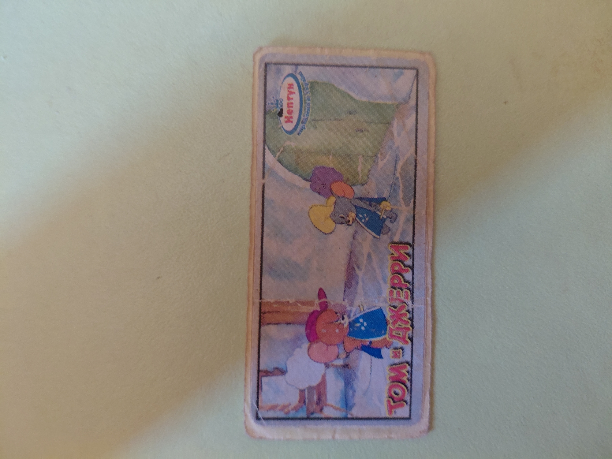 A rectangular sticker depicts cartoon characters Tom and Jerry. The characters are shown in a snowy outdoor scene. On the left is Jerry, wearing a red hat and blue garment. On the right is Tom, wearing a yellow hat and blue garment. In the upper right corner of the sticker is a logo with text that reads "Нептун" and "Мир развлечений и уюта". The sticker is affixed to a light-colored surface.