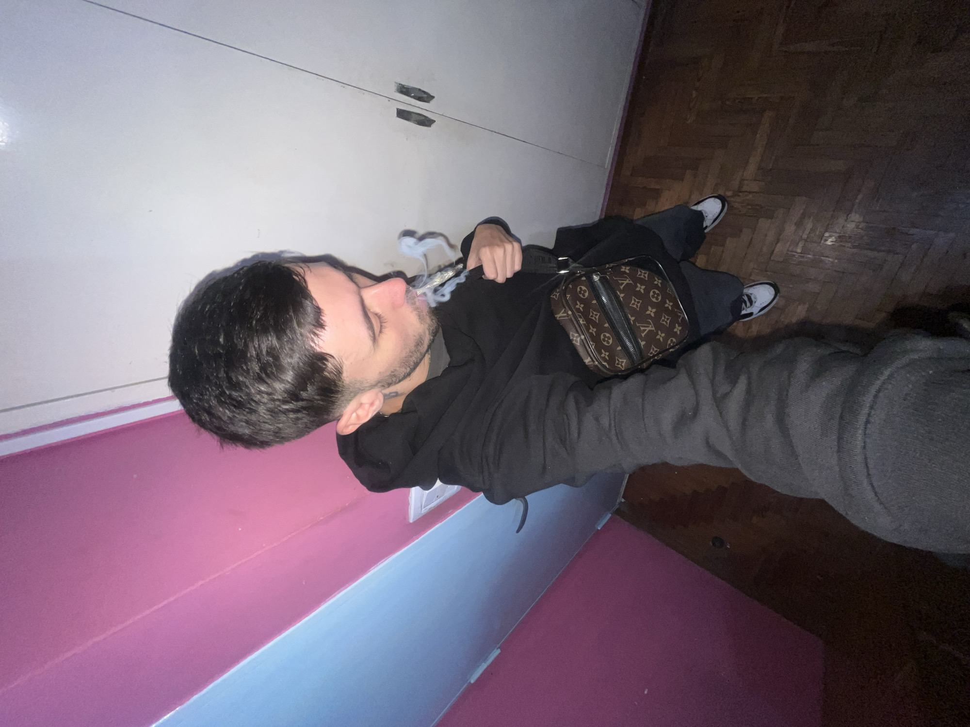 The image depicts a person in a room with pink and light blue walls and a wooden parquet floor. The person is facing the camera, holding what appears to be a lit cigarette or joint, with smoke emanating from it. The person is wearing a black hoodie, dark pants, and black and white sneakers. A brown Louis Vuitton messenger bag is slung across their body. Behind them, there's a white cabinet with metal handles. The location is specified as Llavallol, Argentina, but there is no additional data that allows for forensic information about this city.