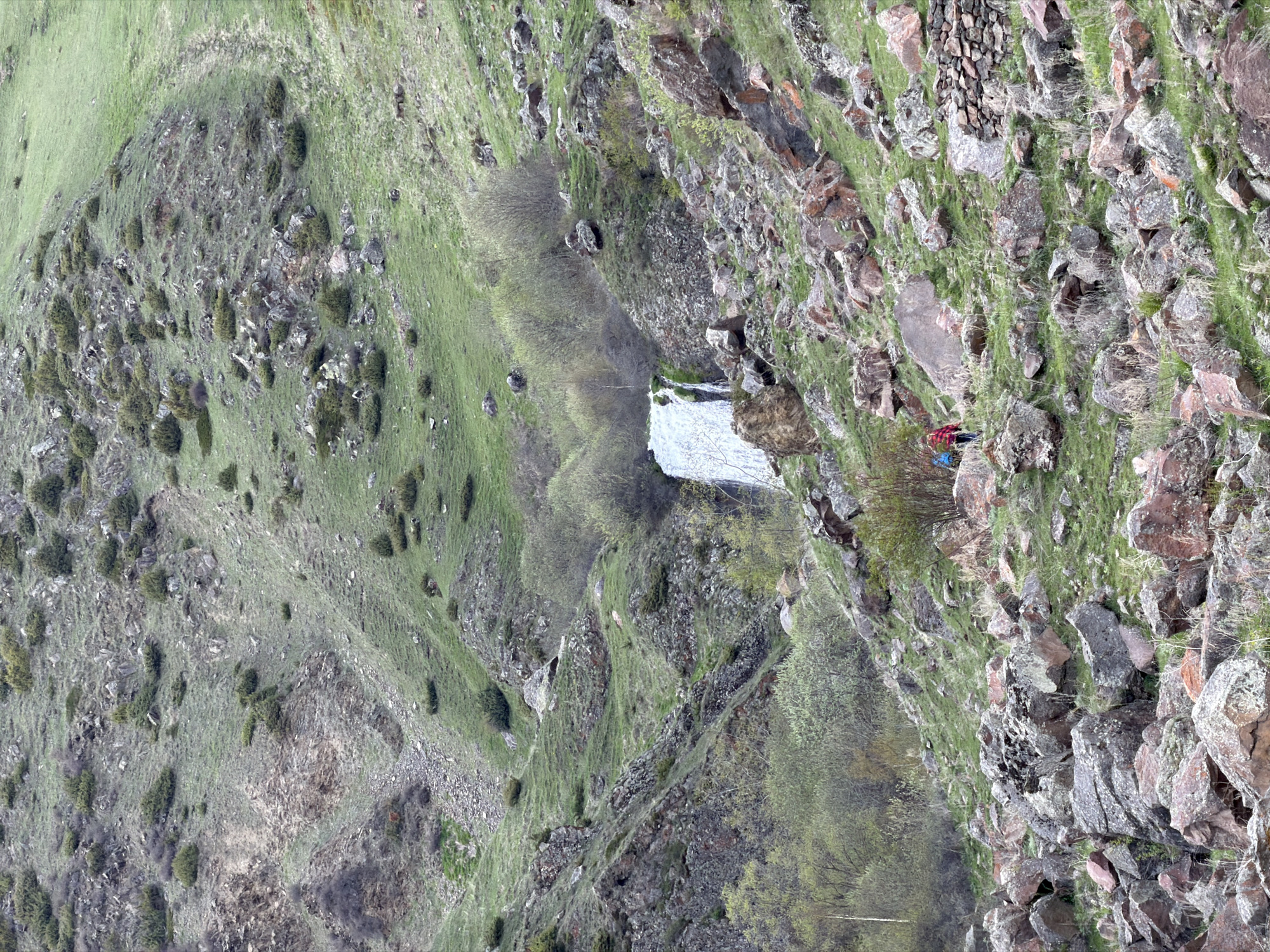 The image depicts a rocky, mountainous landscape in Arsha, Georgia. The terrain consists of steep slopes with patches of grass and sparse vegetation. There's a waterfall visible amidst the rocks and trees. A person is positioned on the right side of the image on a rocky outcrop. They appear to be wearing a red jacket and a blue backpack. The overall environment seems natural, with a rugged, untouched quality.